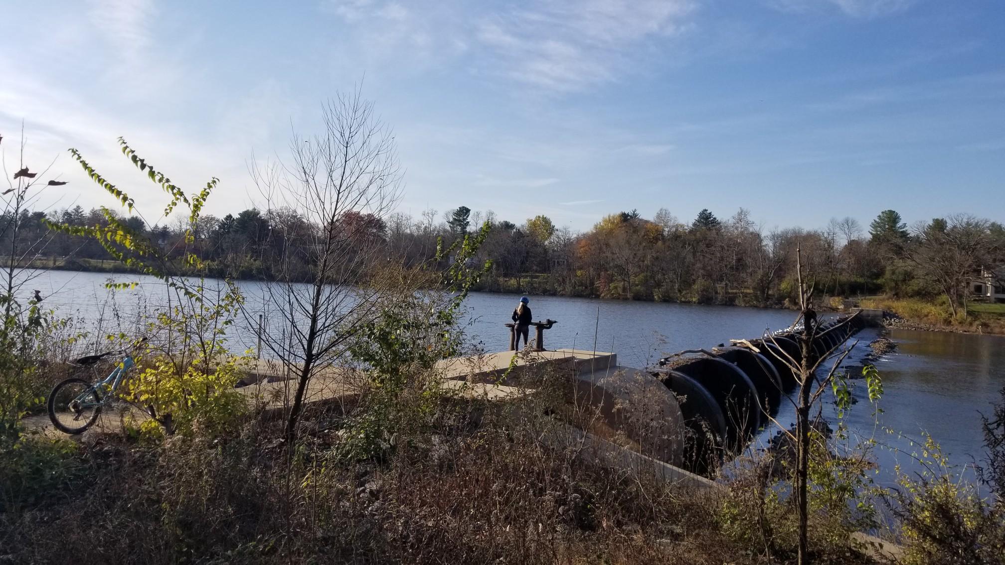 A person fishes by a lake with a dam under a bright blue sky.