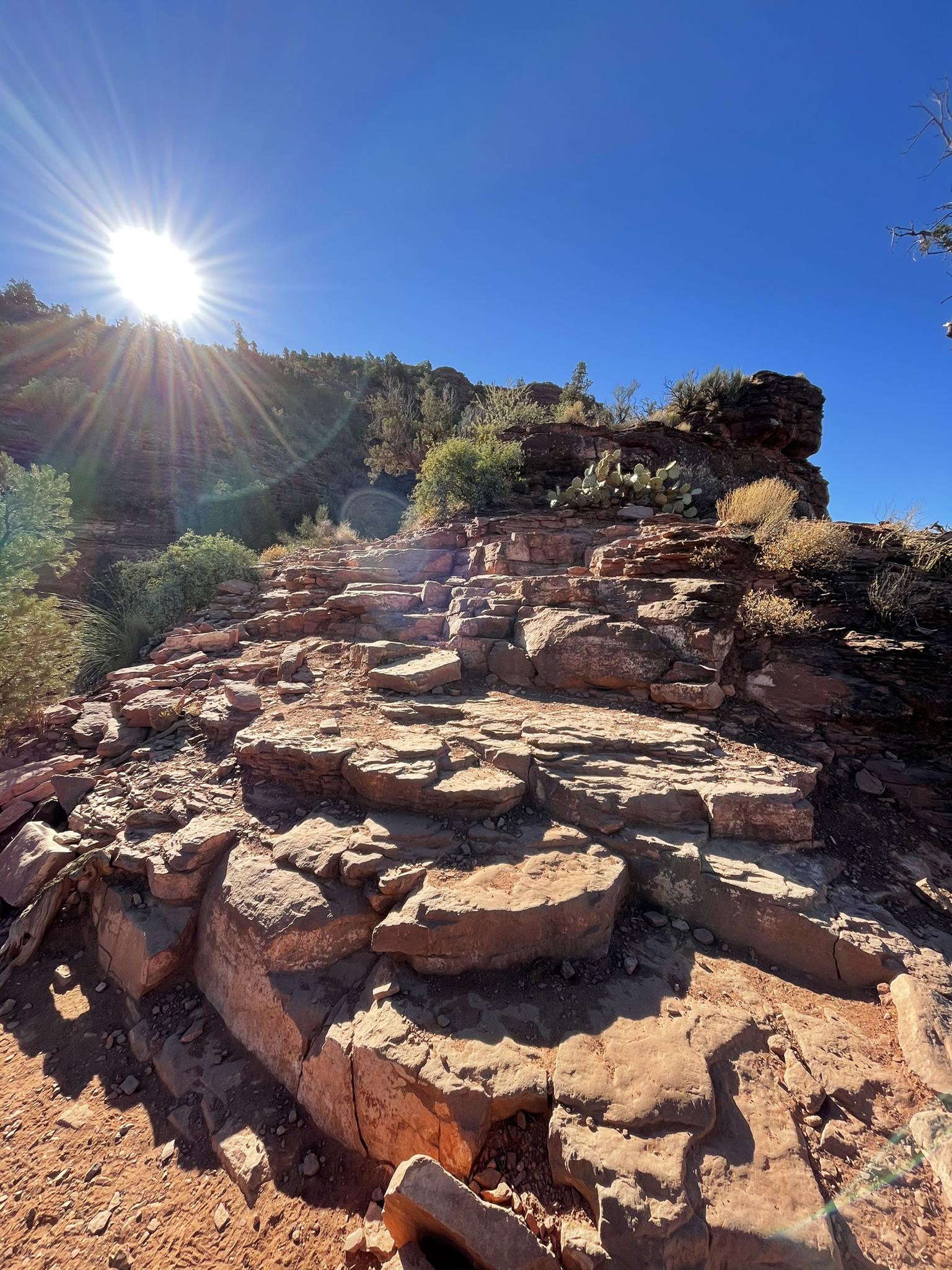 Bright sunburst over rocky red terrain with steps and desert plants.