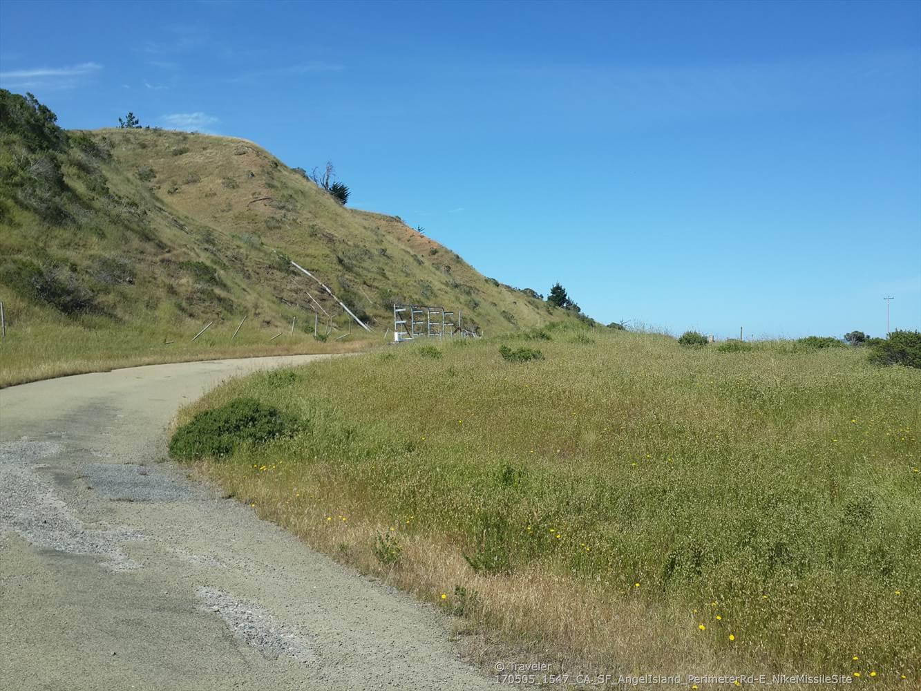 Winding dirt road through green and golden grassy hills under blue sky.