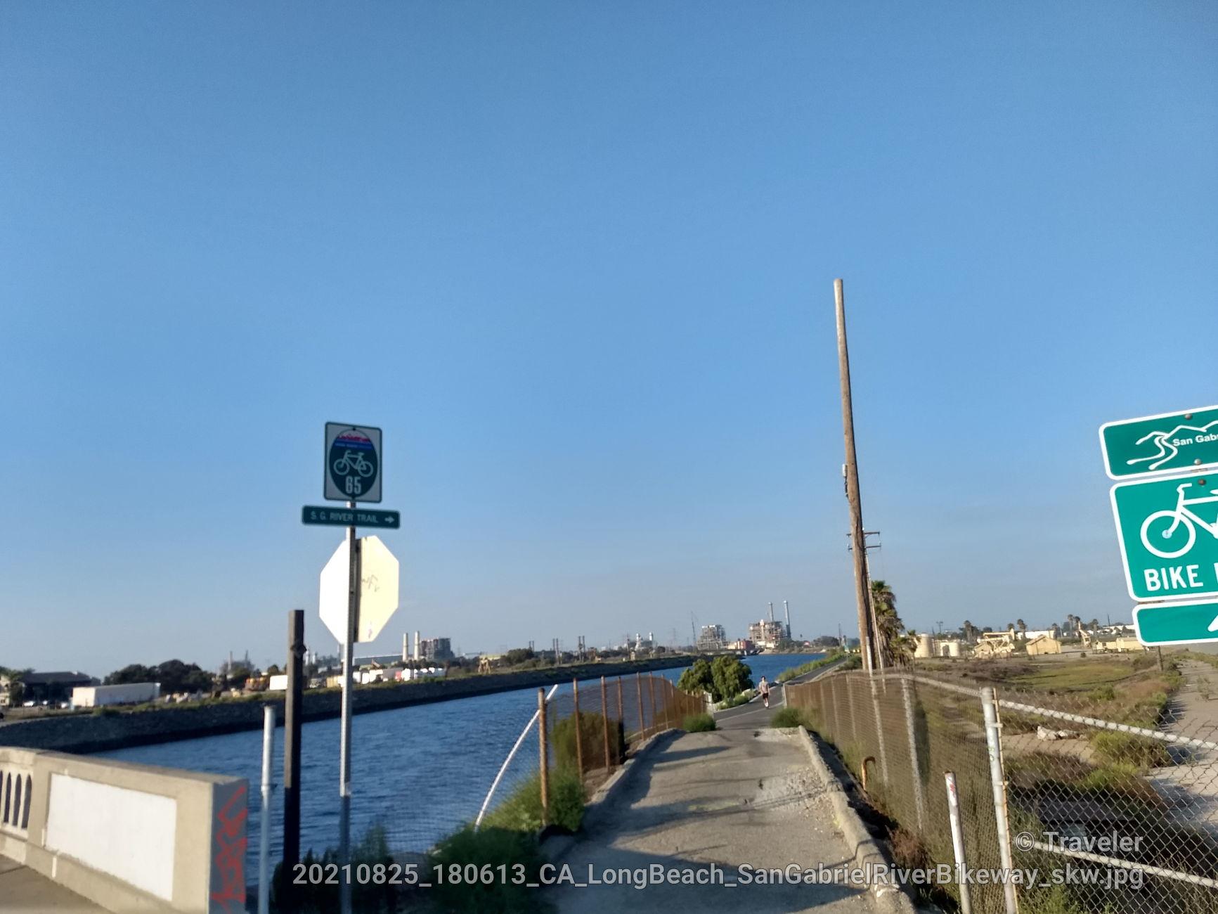A paved path next to a canal under a clear blue sky with distant buildings.
