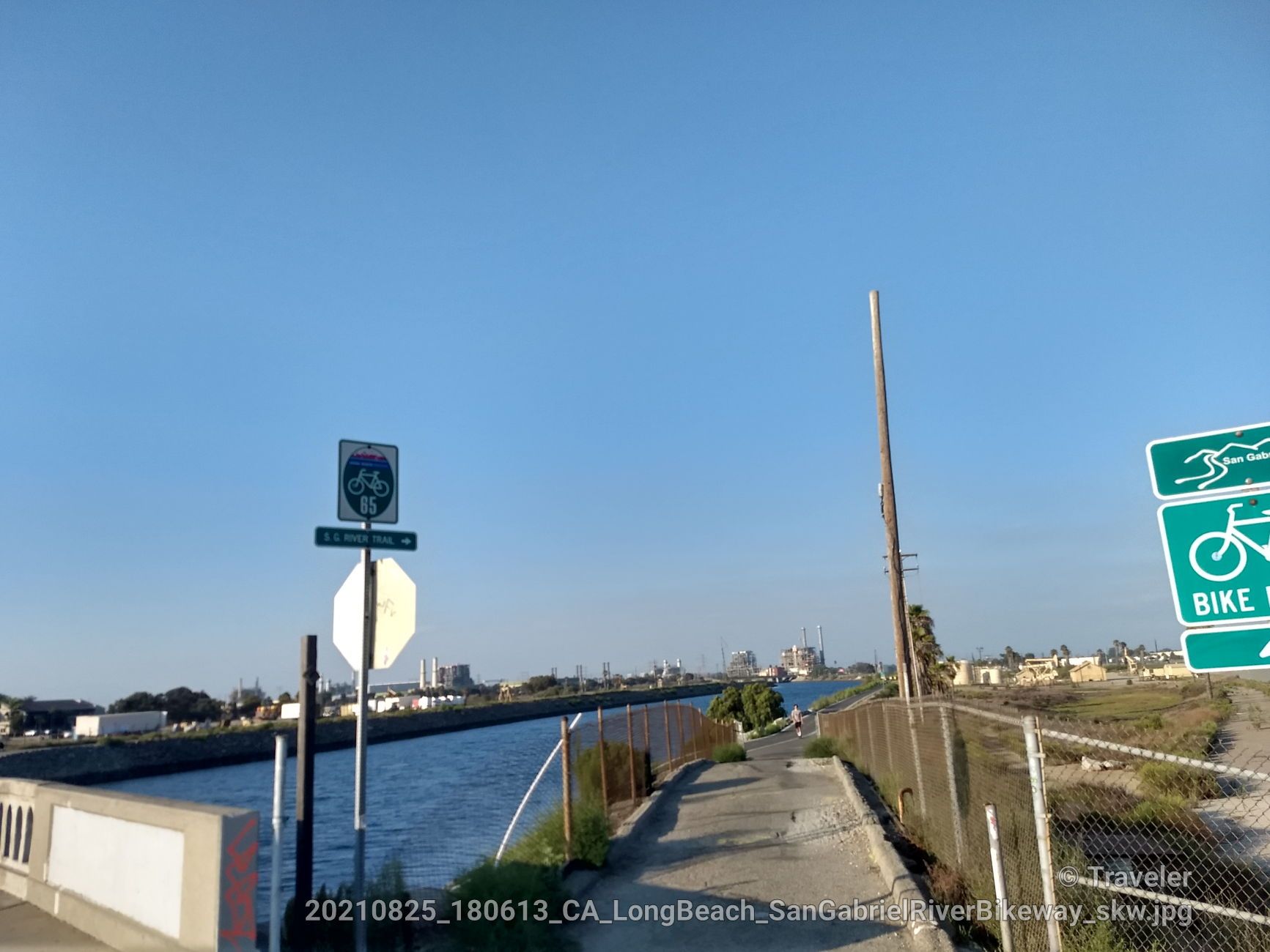 A paved path next to a canal under a clear blue sky with distant buildings. - San Gabriel River Trail