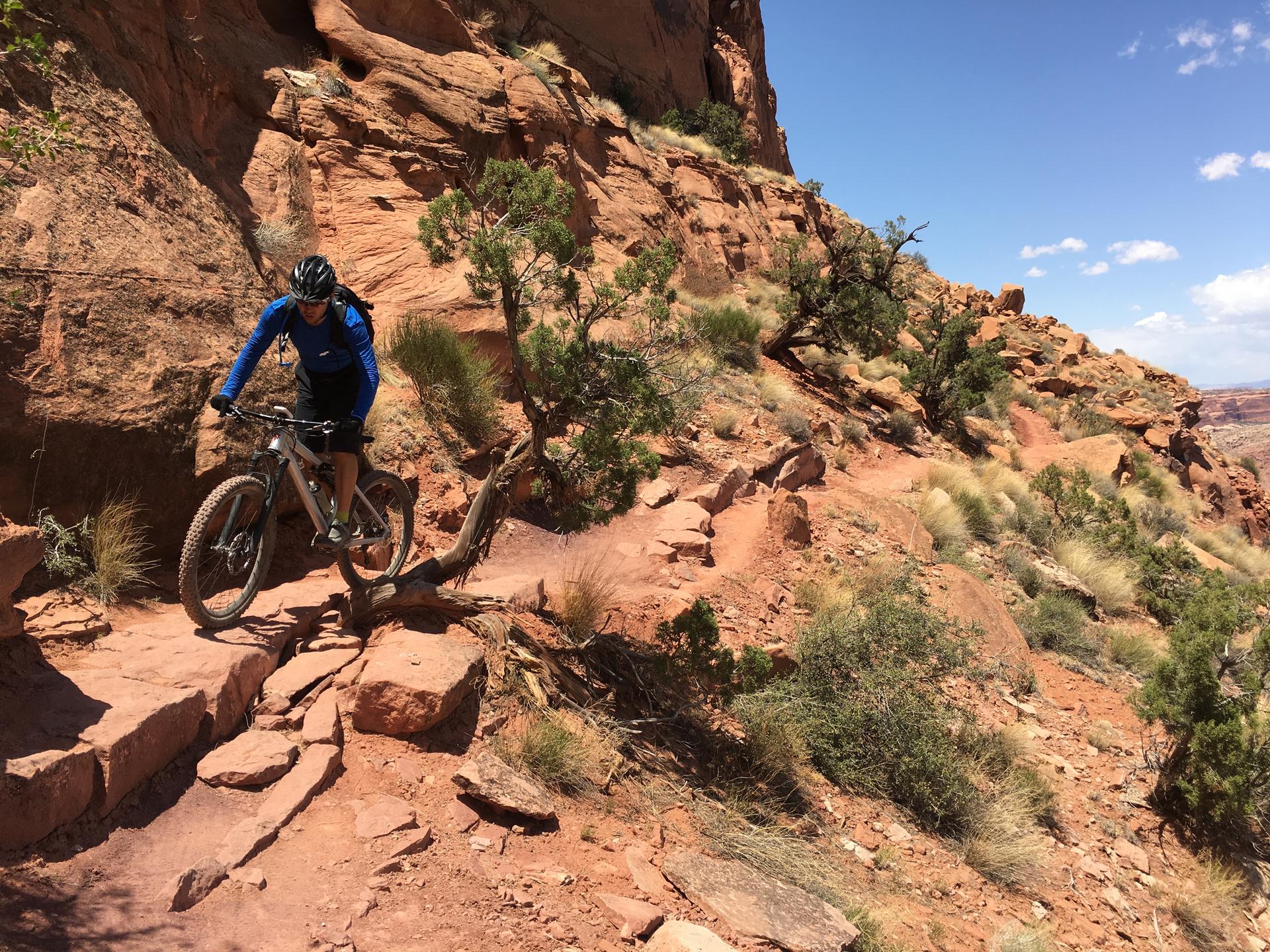 Mountain biker navigates a steep, rocky trail on a sunny day with red cliffs.