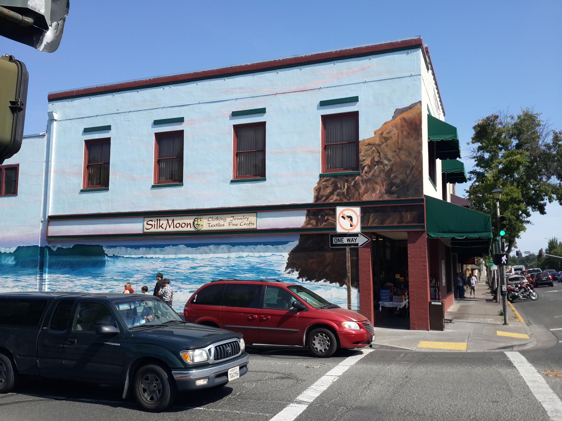 Building with a large mural on a sunny street corner with parked cars.