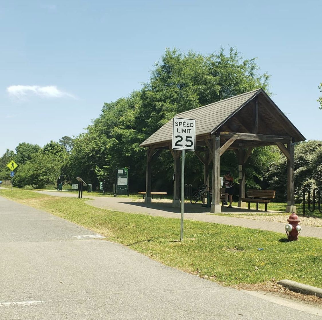 Wooden shelter with people resting, next to a paved path on a sunny day. - Virginia Capital Trail