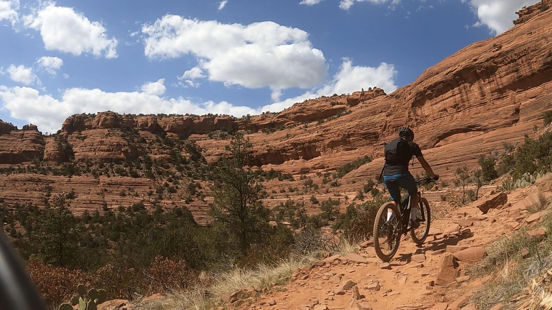 Mountain biker descends a red dirt trail beside towering red rock cliffs.