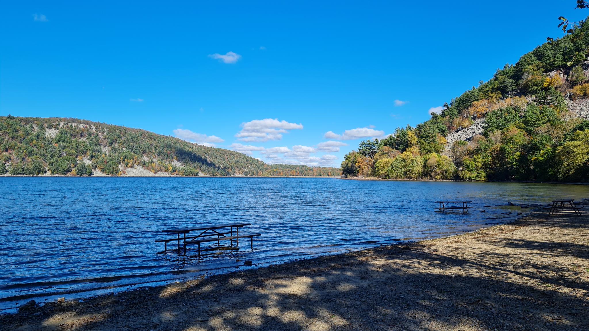 Sunny day at a lake with forested hills and submerged picnic tables.