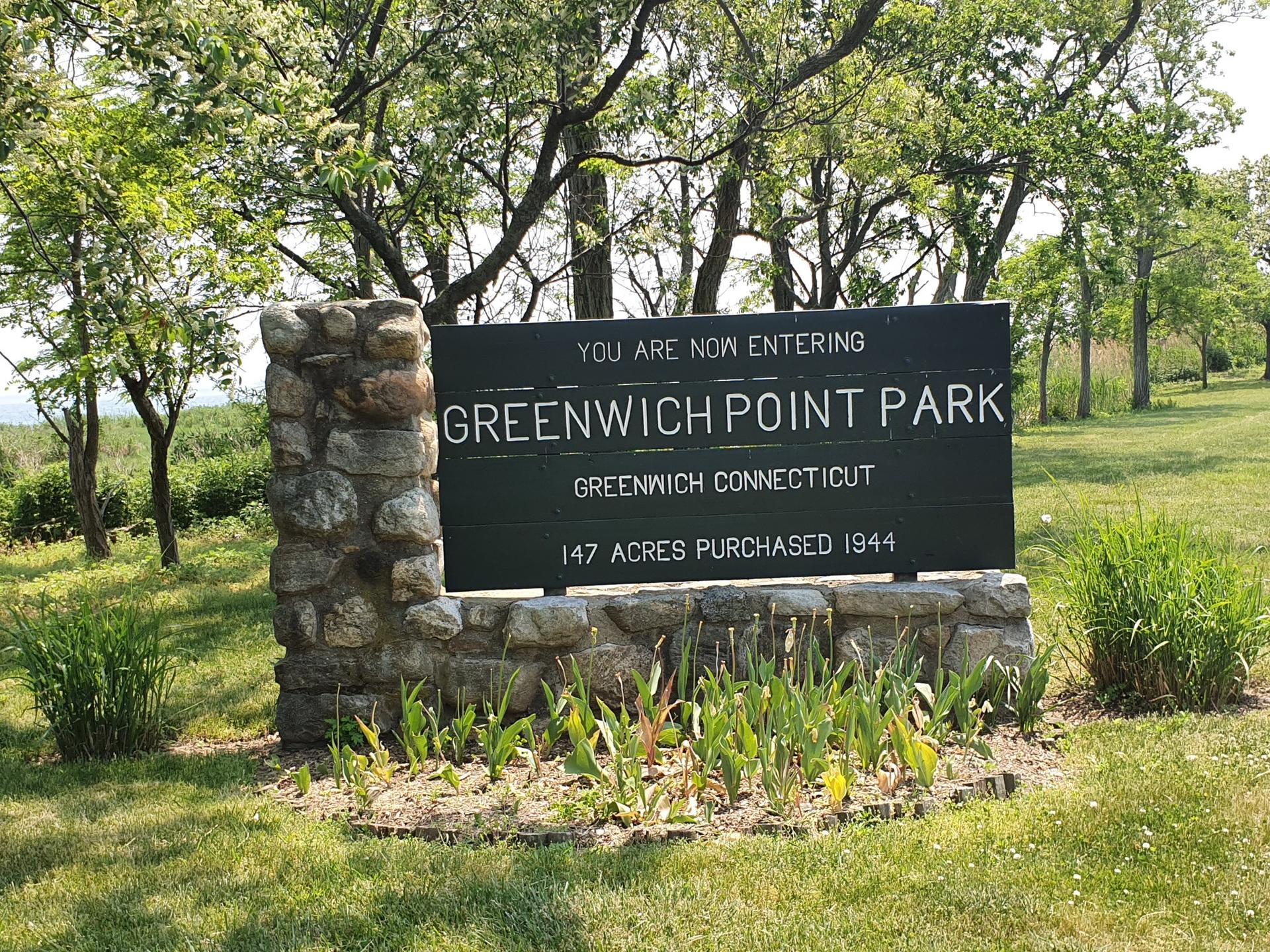 A large dark sign on a stone base in a sunny, grassy park.