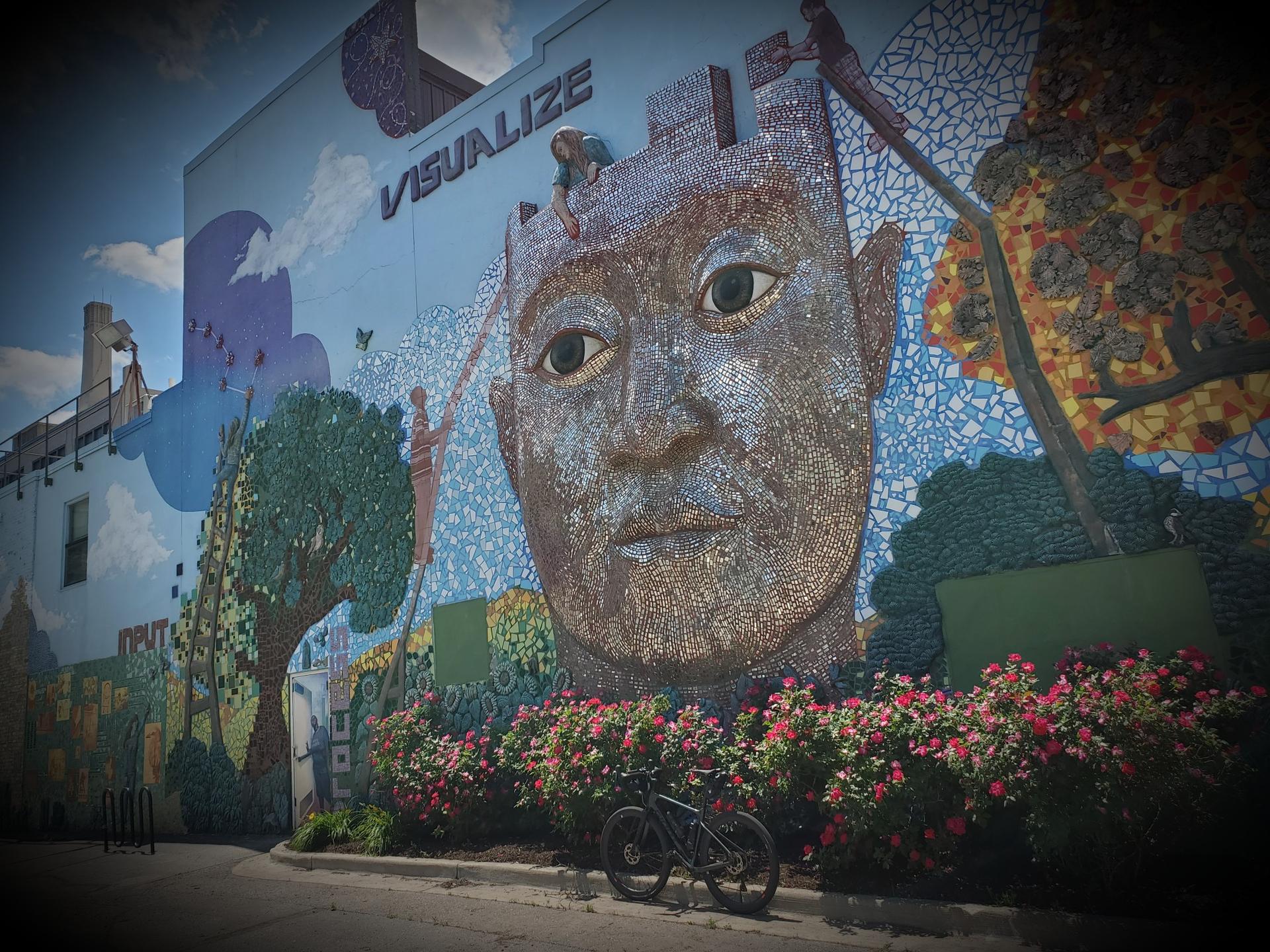 Large mosaic mural of a man's face on a building, with a bicycle.