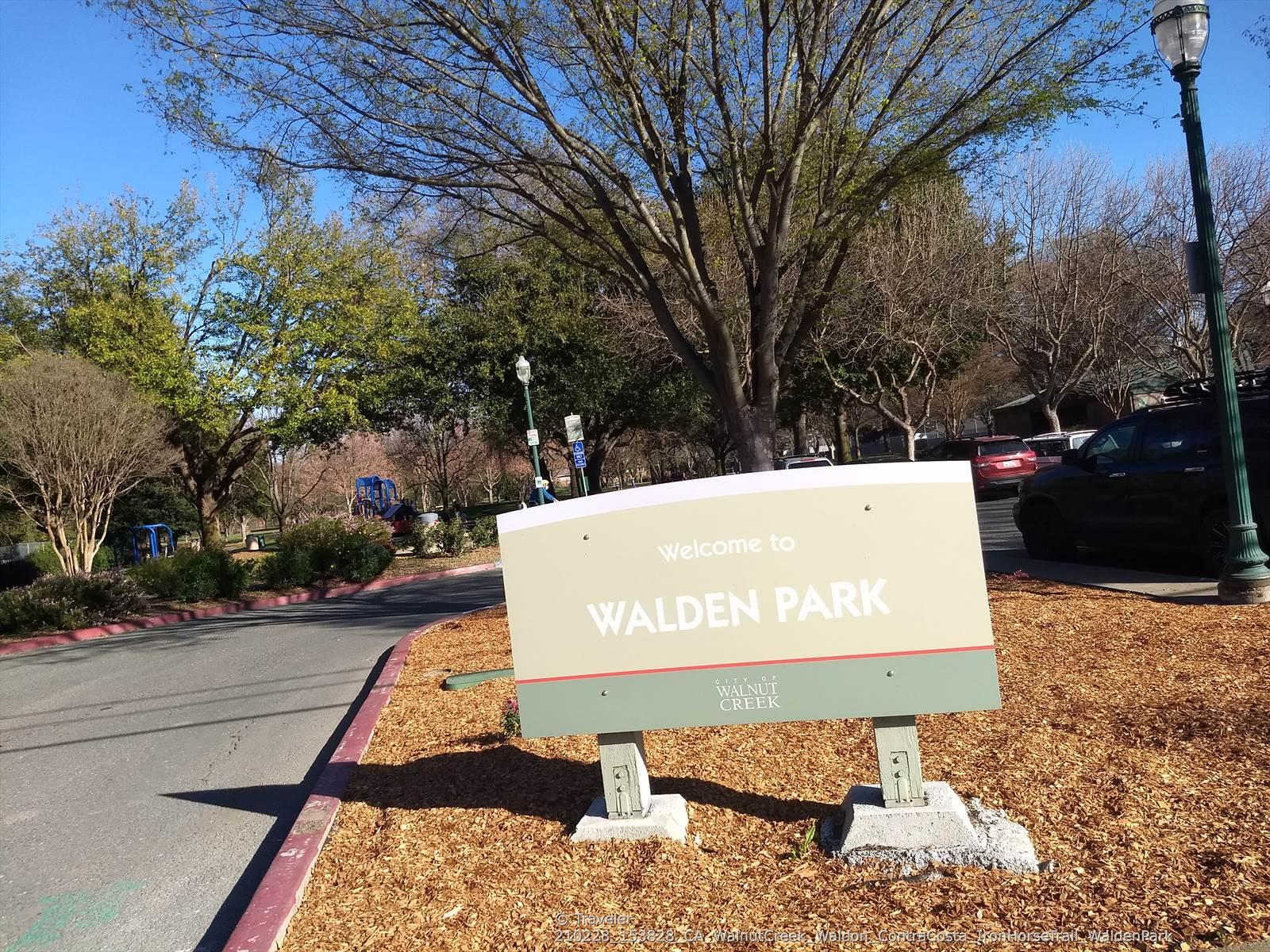 A park sign on a sunny day, surrounded by trees and dry mulch.