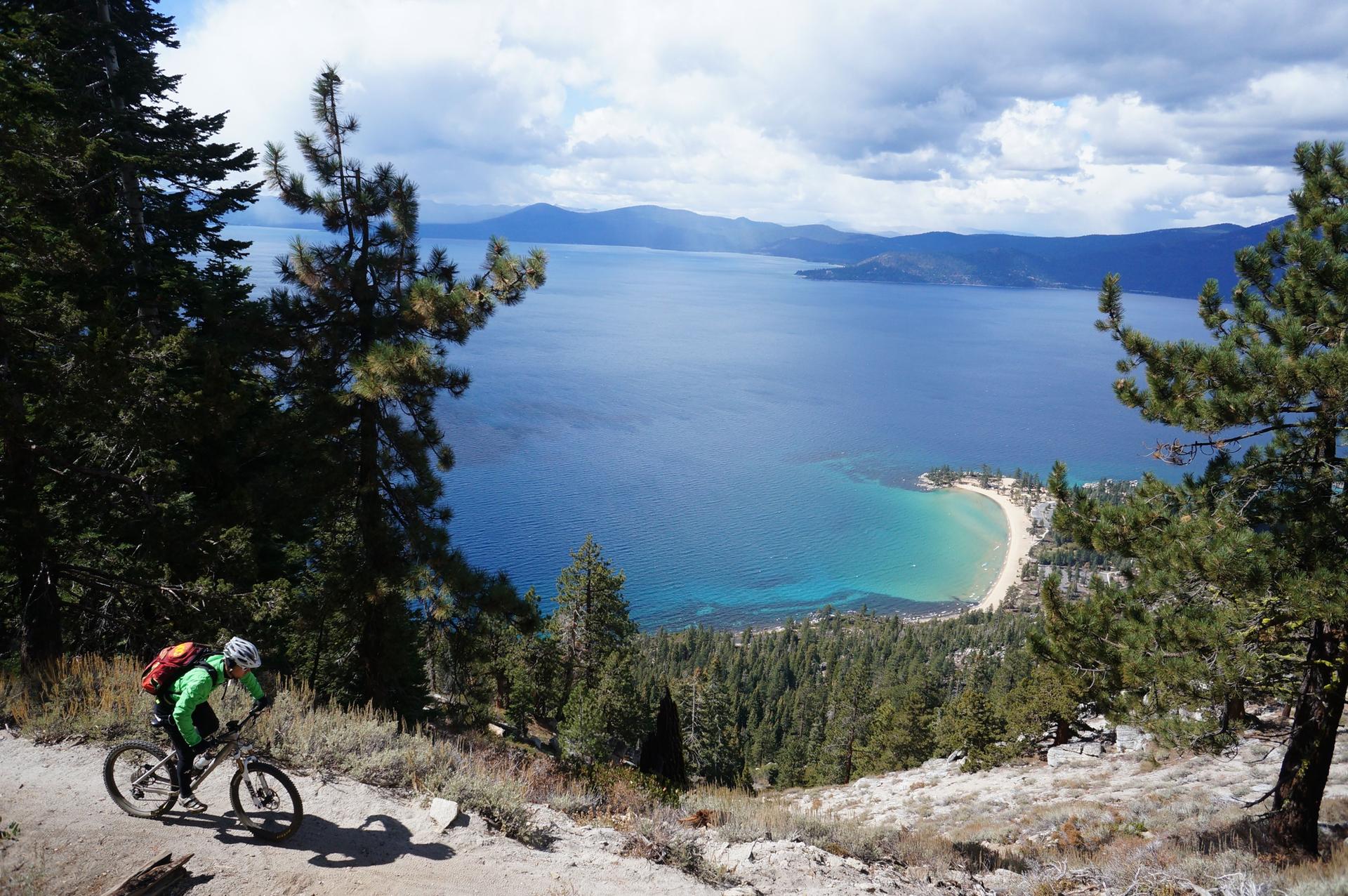 Mountain biker on a dirt trail overlooking a vast blue lake and forested mountains.