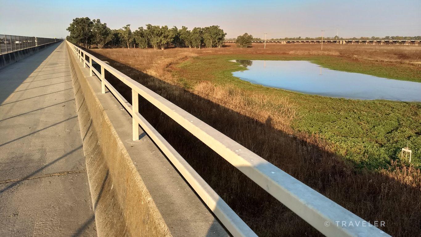 A concrete bridge railing overlooks a pond and golden fields under a clear sky.