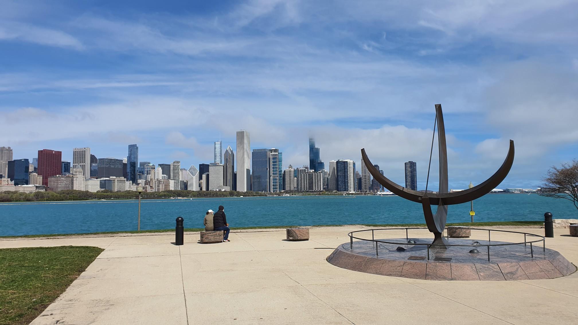 People near a large sundial sculpture with a city skyline across water.