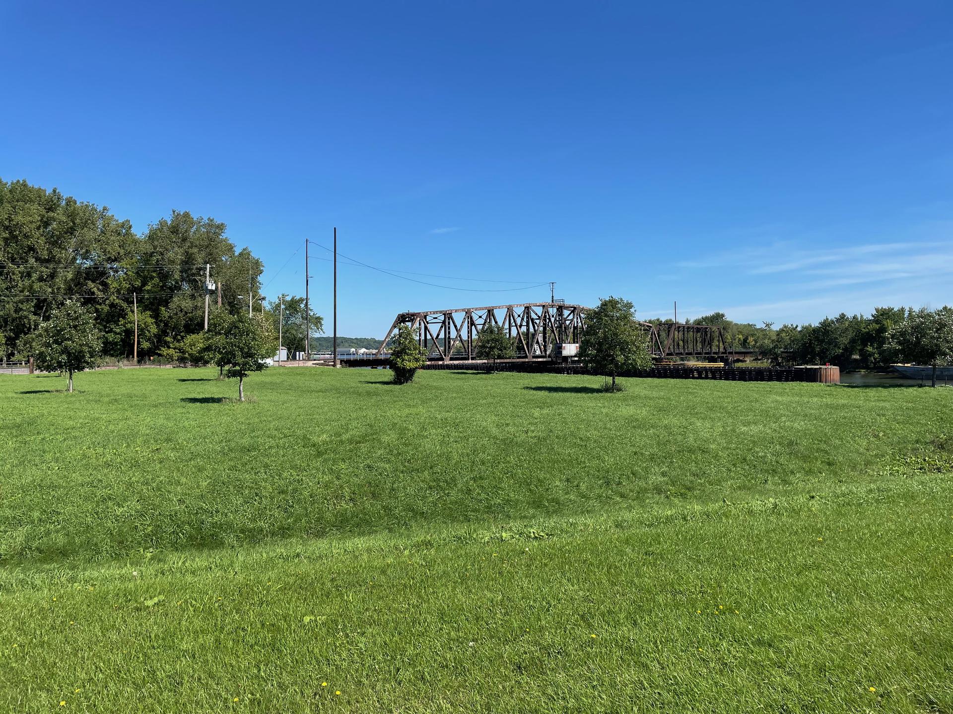 Expansive green field with a distant metal bridge under a clear blue sky.