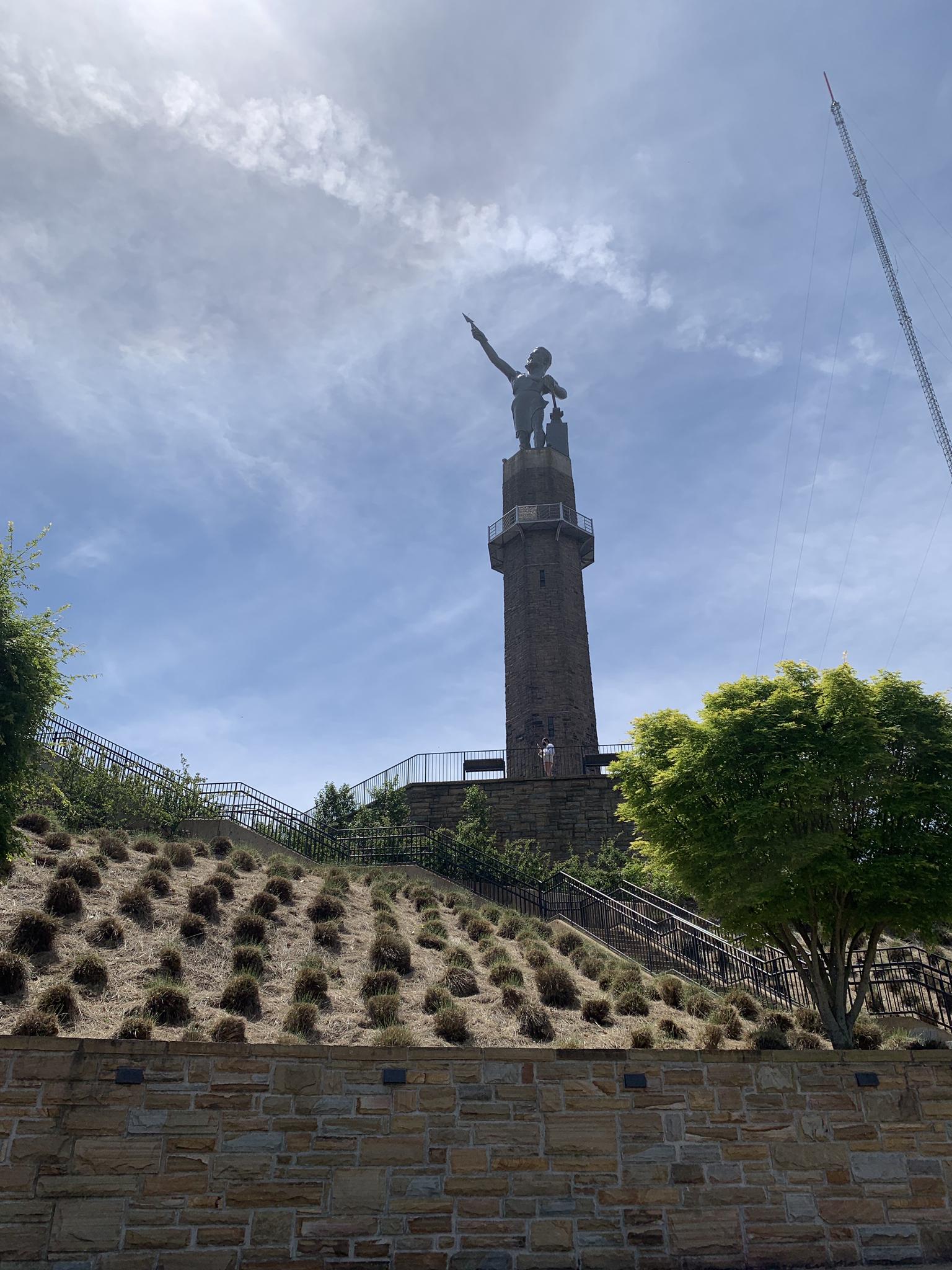 Tall statue on a tower pointing skyward, with a hillside and stone wall below.