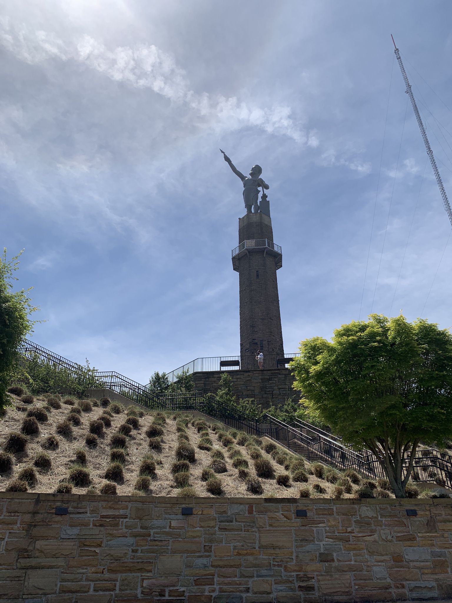 Tall statue on a tower pointing skyward, with a hillside and stone wall below. - Kiwanis Vulcan Trail