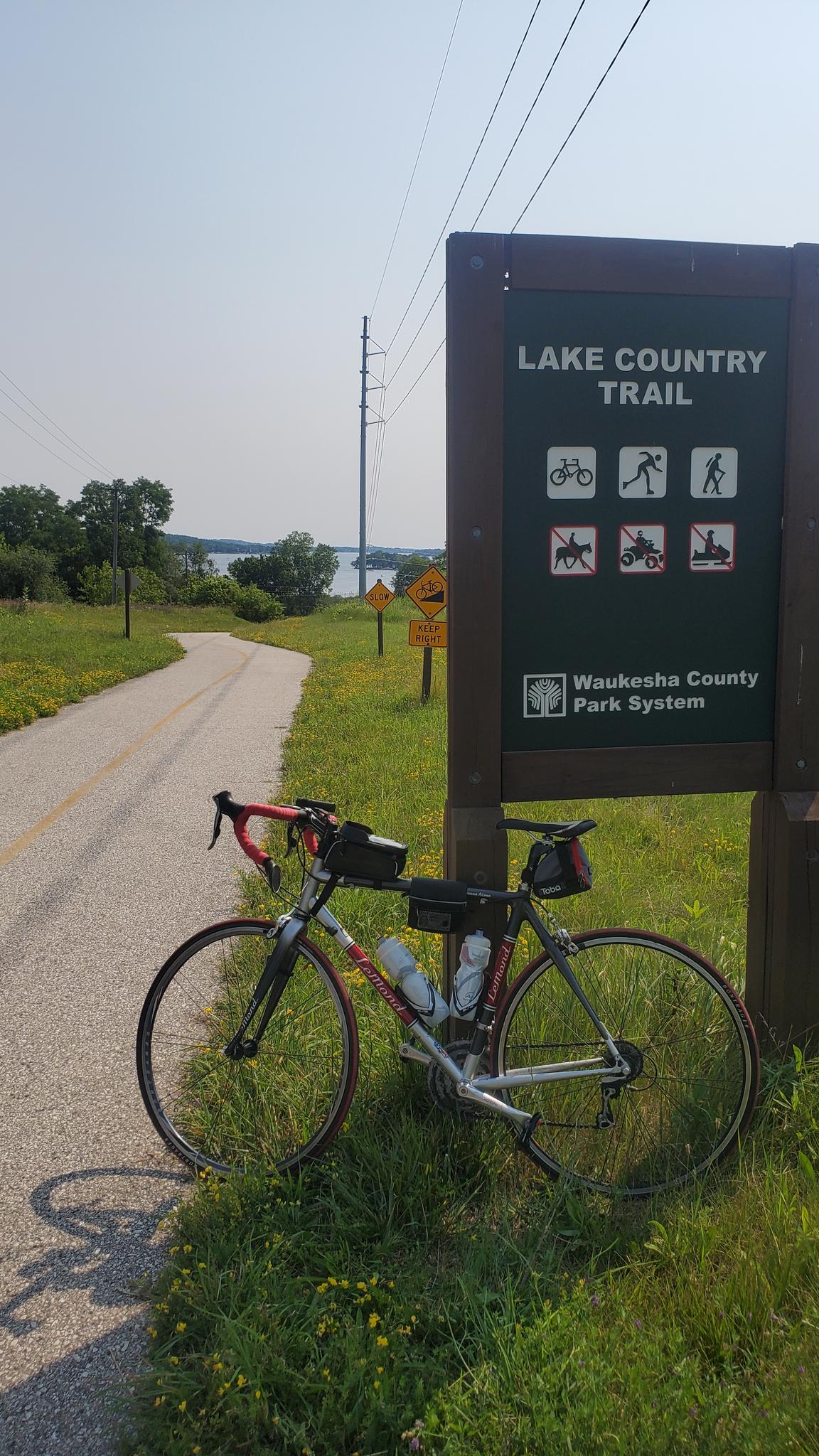 Bicycle parked next to a trail sign on a sunny day by a lake.