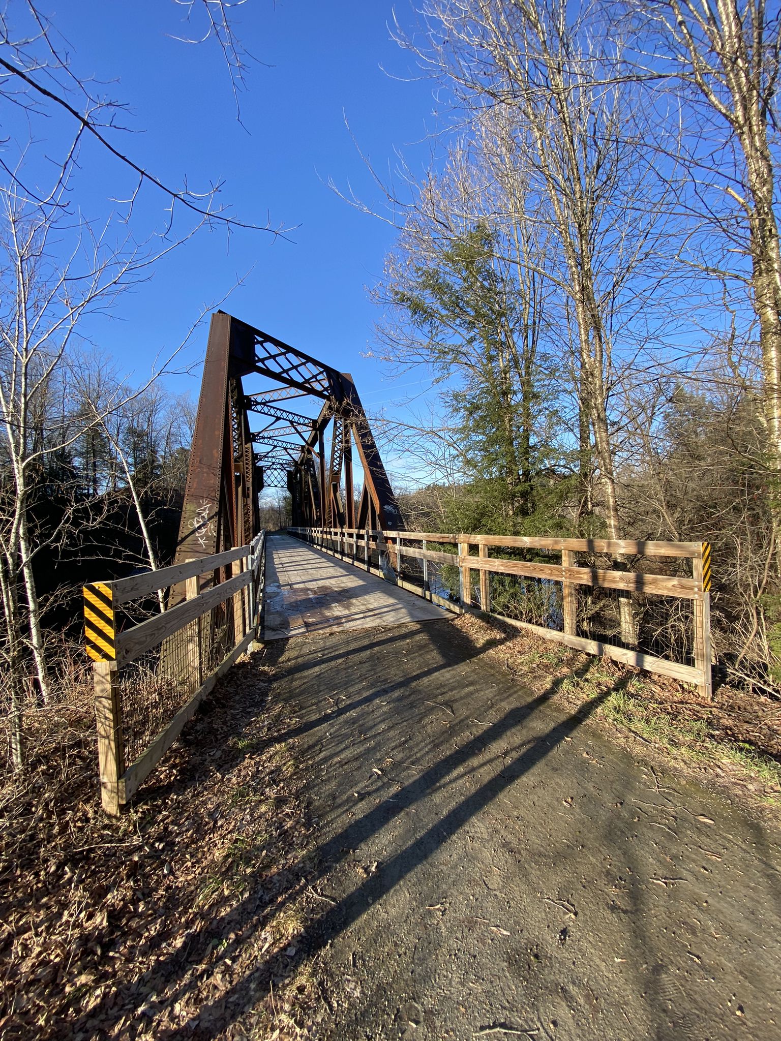 A metal truss bridge spans a path with bare trees under a blue sky. - Old Railroad Bridge – Dog’s Head Falls loop from Morristown