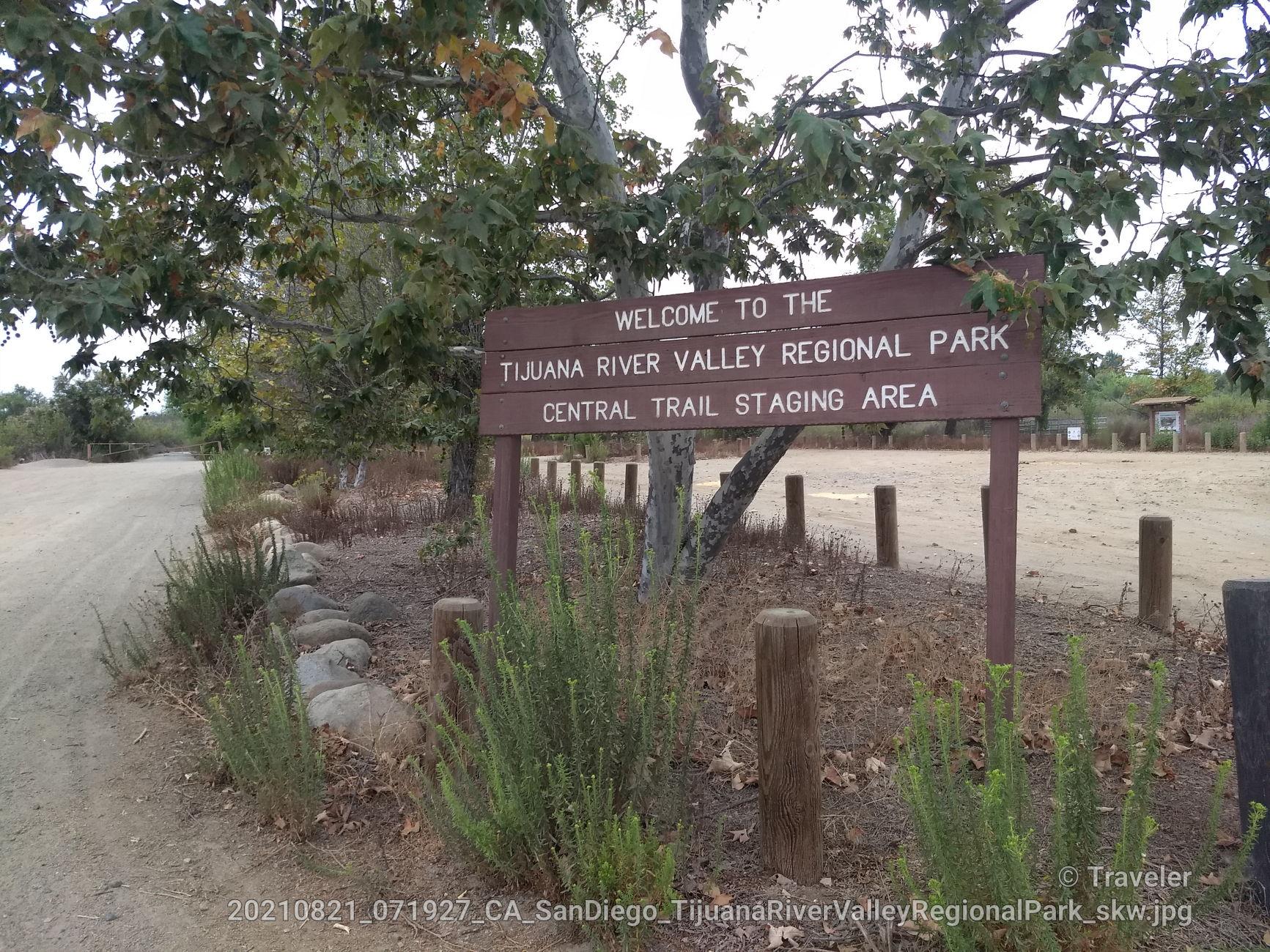 A brown wooden sign stands beside a dirt path, surrounded by green trees.