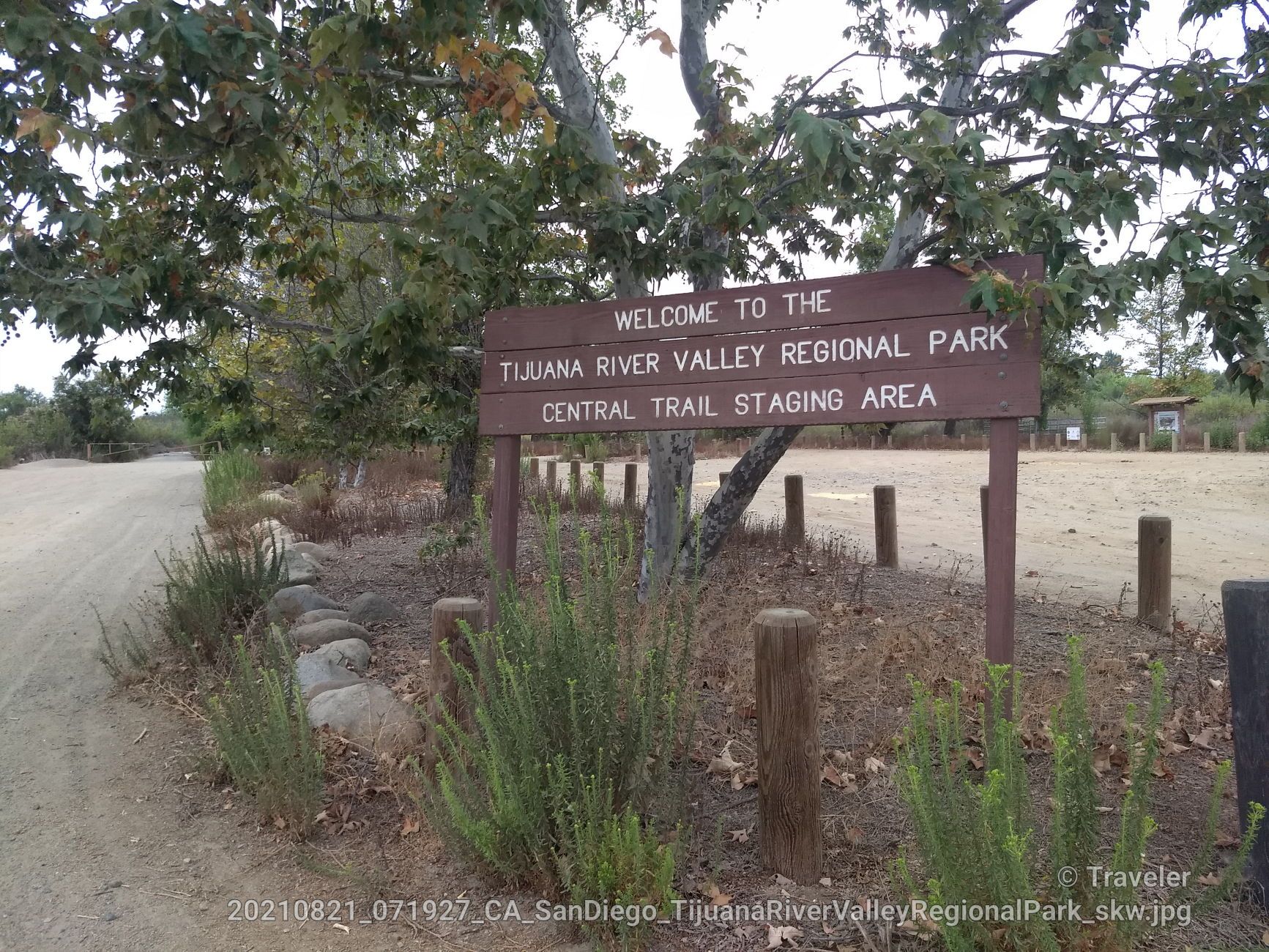 A brown wooden sign stands beside a dirt path, surrounded by green trees. - Olympic Parkway to Tijuana River Valley Regional Park loop