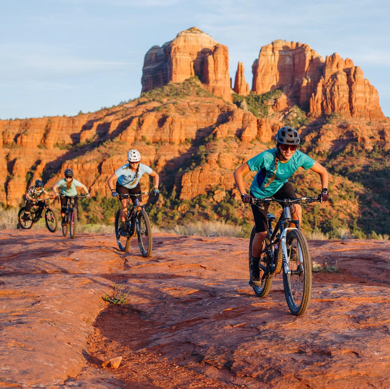 Four mountain bikers ride on red rock trails with large rock formations at sunset.