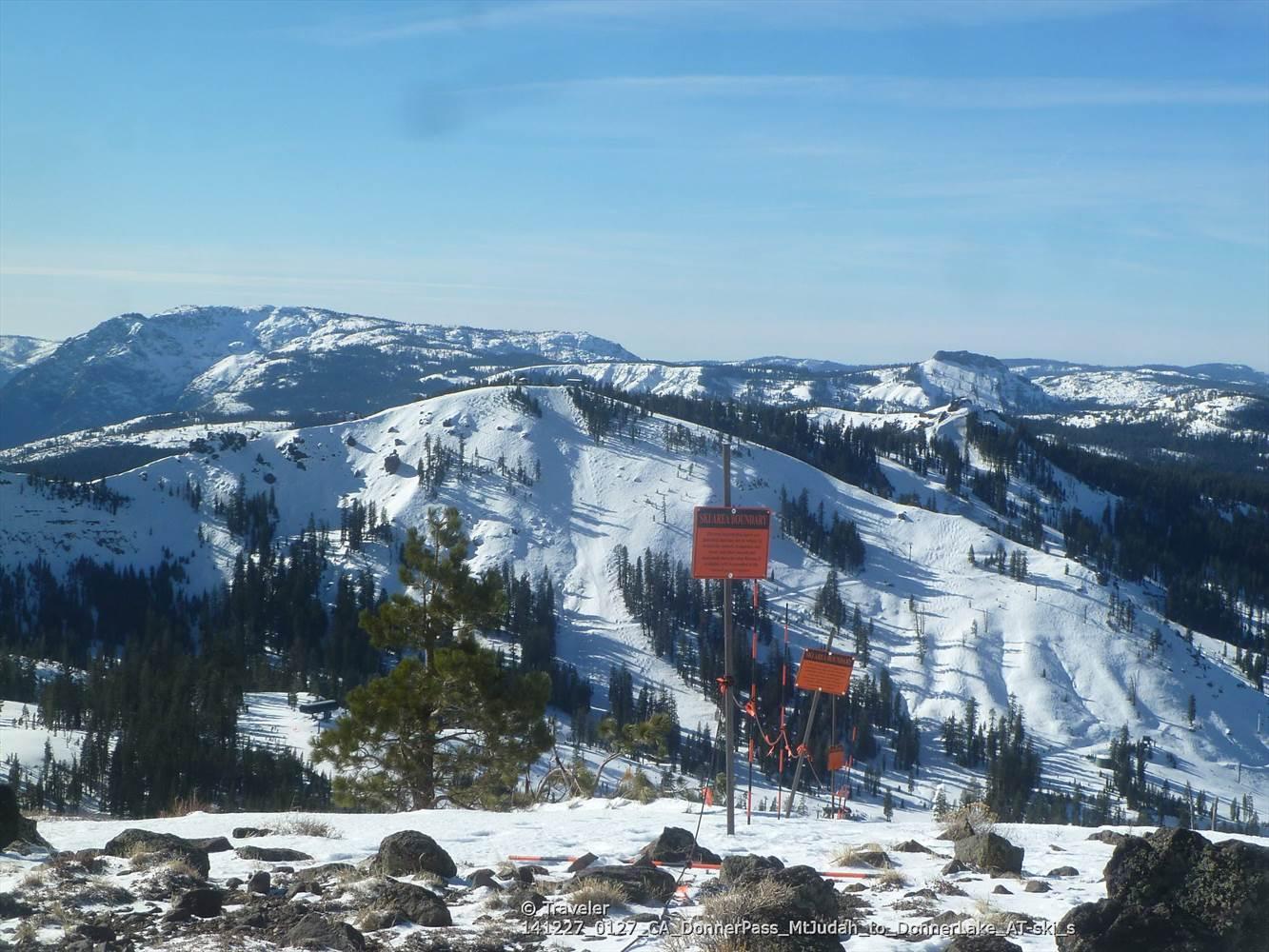 Snow-covered mountains and pine trees under a clear blue sky, with orange ski markers.