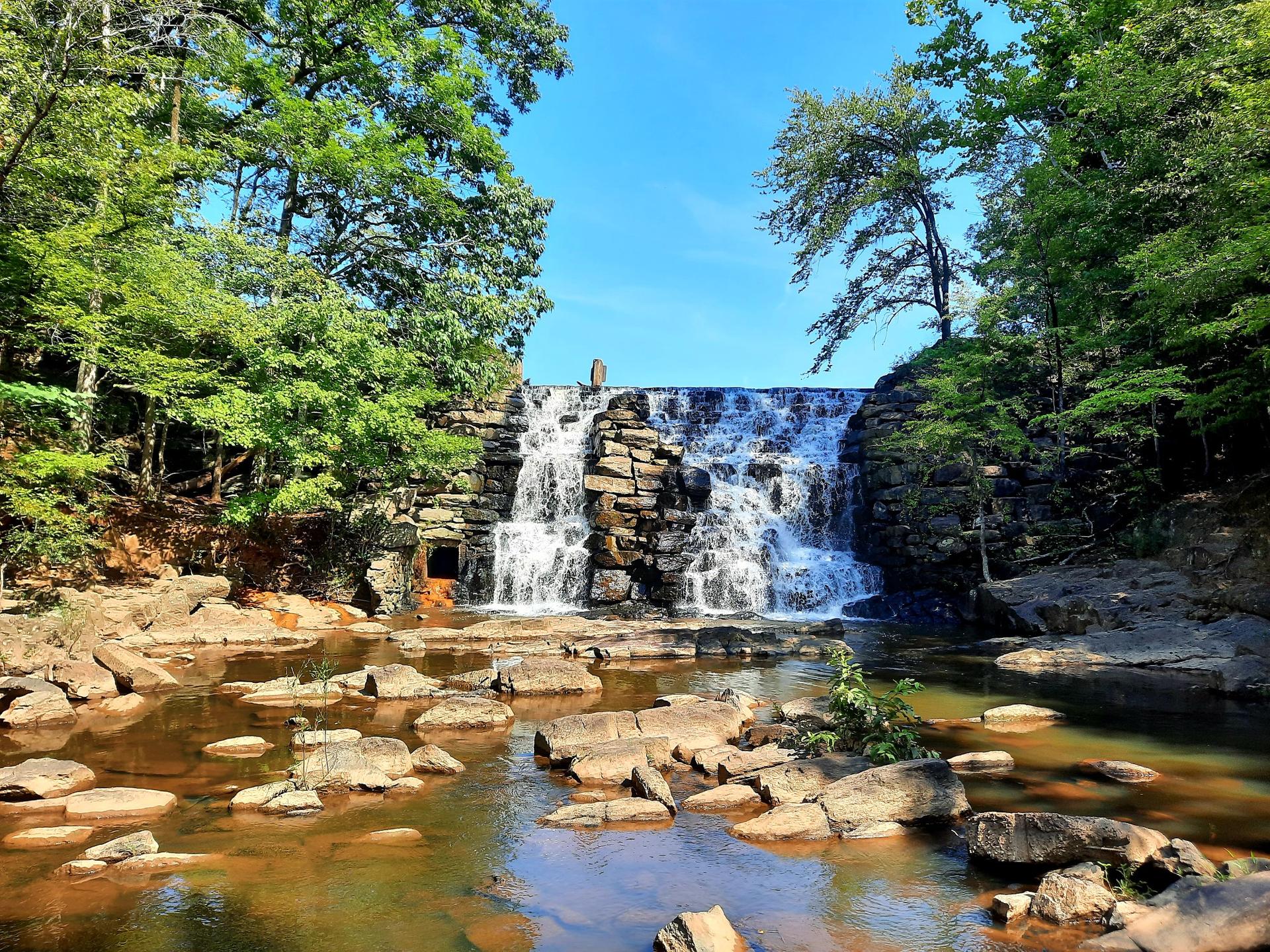 Waterfall over a stone dam, rocky river in foreground, bright sunny day.