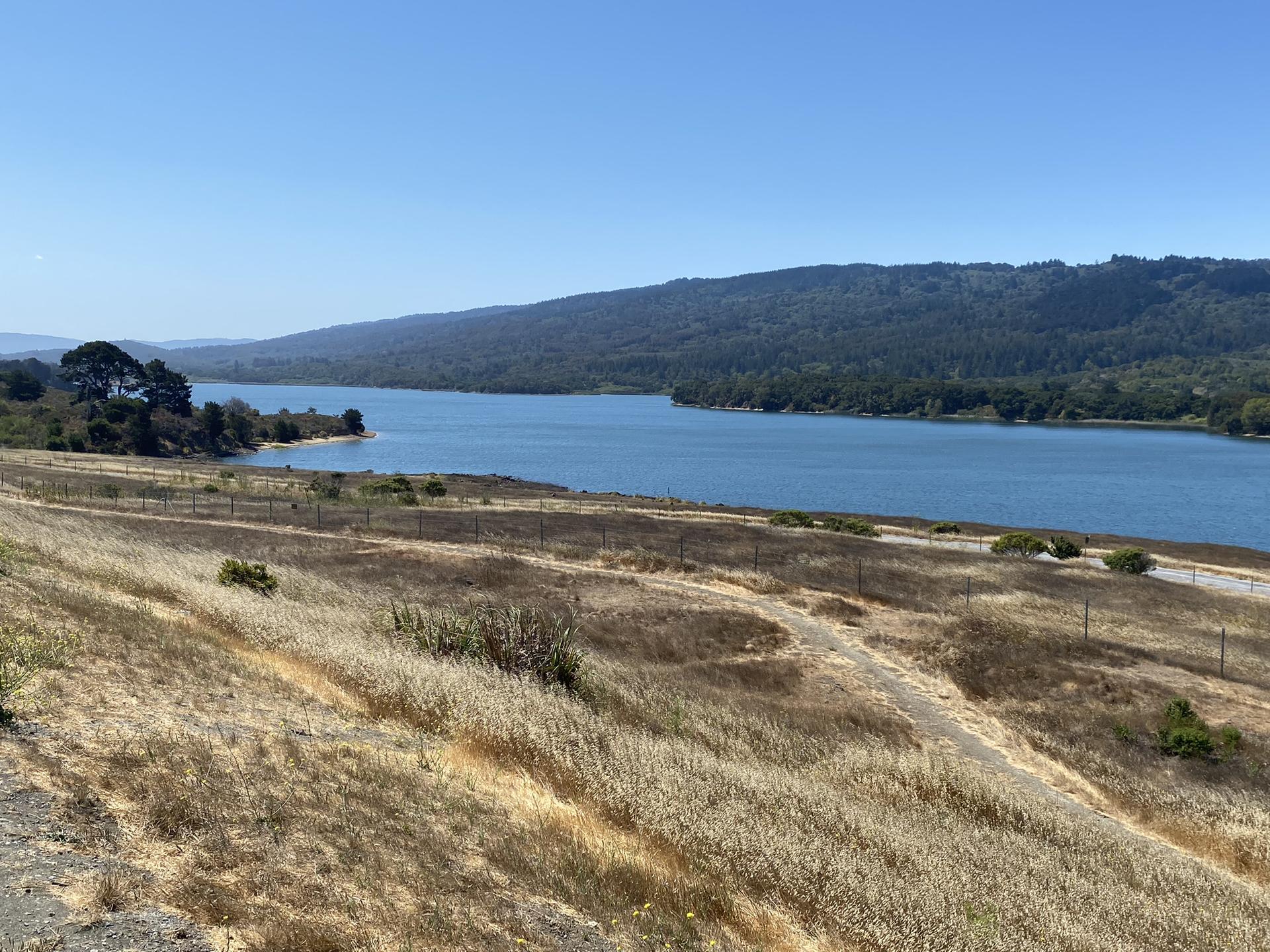 Sunny view of a large lake bordered by dry grass and forested hills.