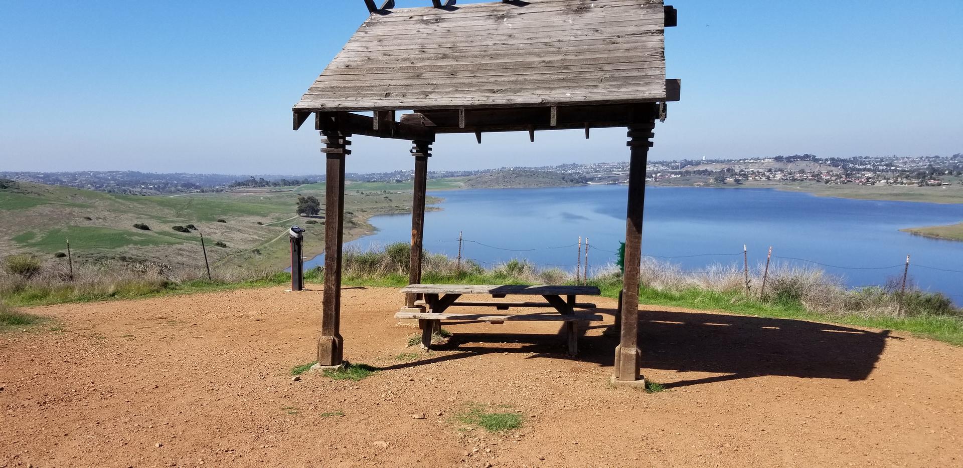 Wooden gazebo and picnic table overlooking a vast blue lake on a sunny day.