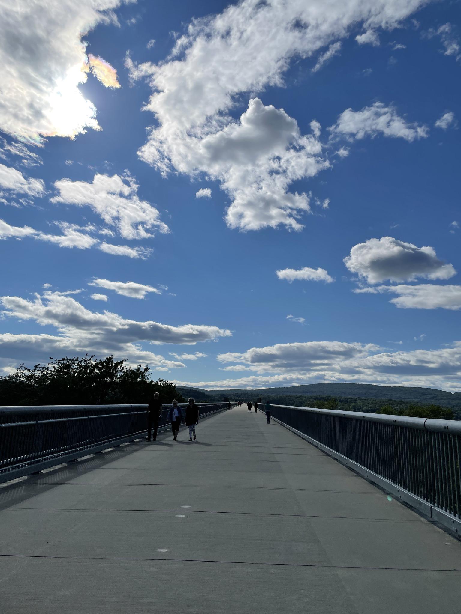 People walking on a long, elevated pathway under a bright, cloudy sky.