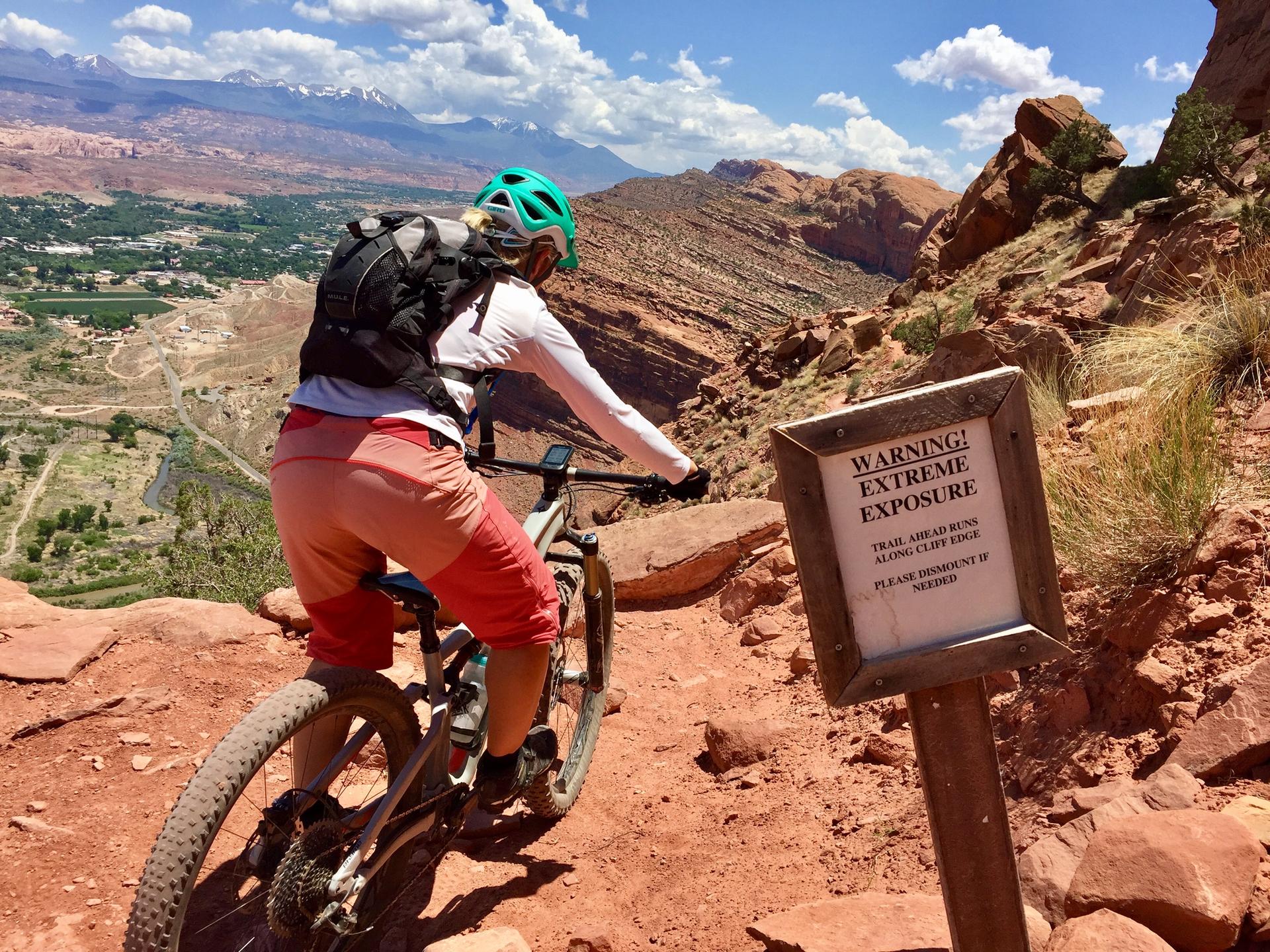 Mountain biker on a narrow red rock trail with a warning sign and vast canyon views.