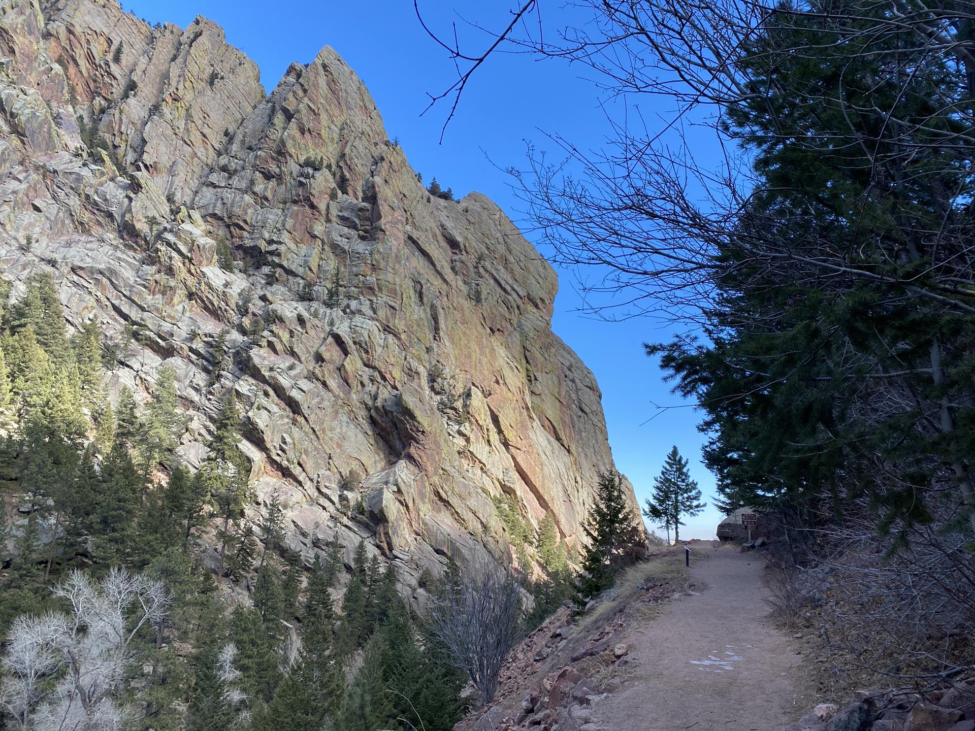 Steep rock face beside a dirt path with pine trees under a clear sky.