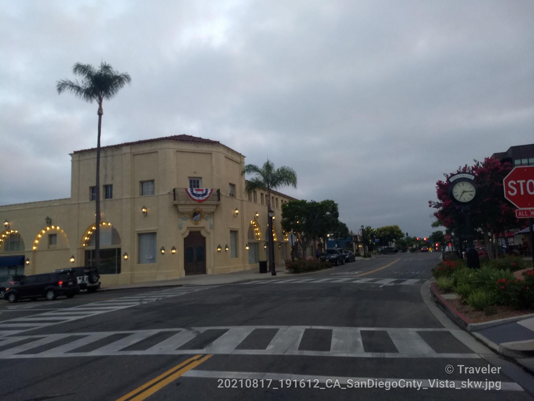 Street with a large building, palm trees, and a stop sign under cloudy skies.