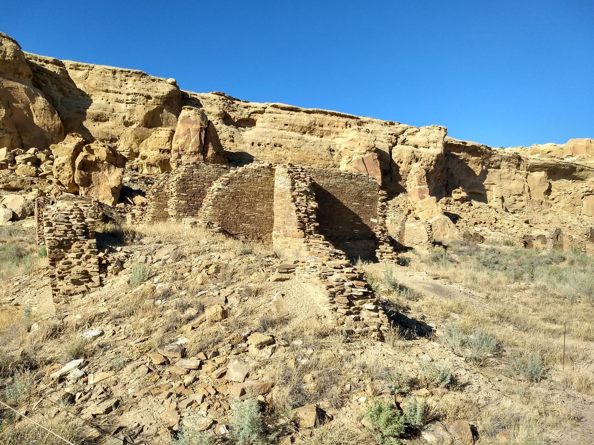 Stone ruins against a rocky cliff face, bright desert sunlight.