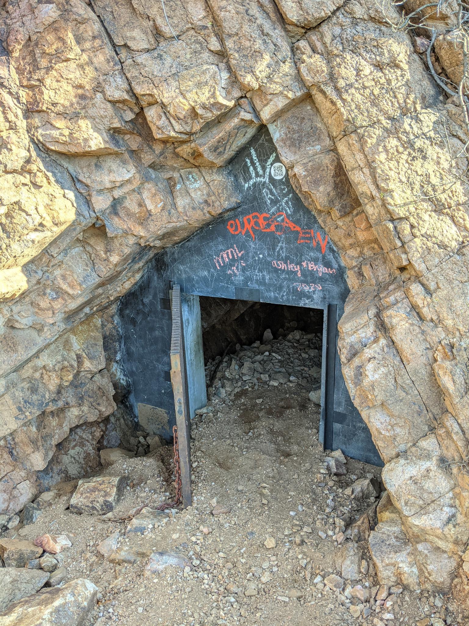 Dark mine entrance carved into a rocky cliff, with graffiti.