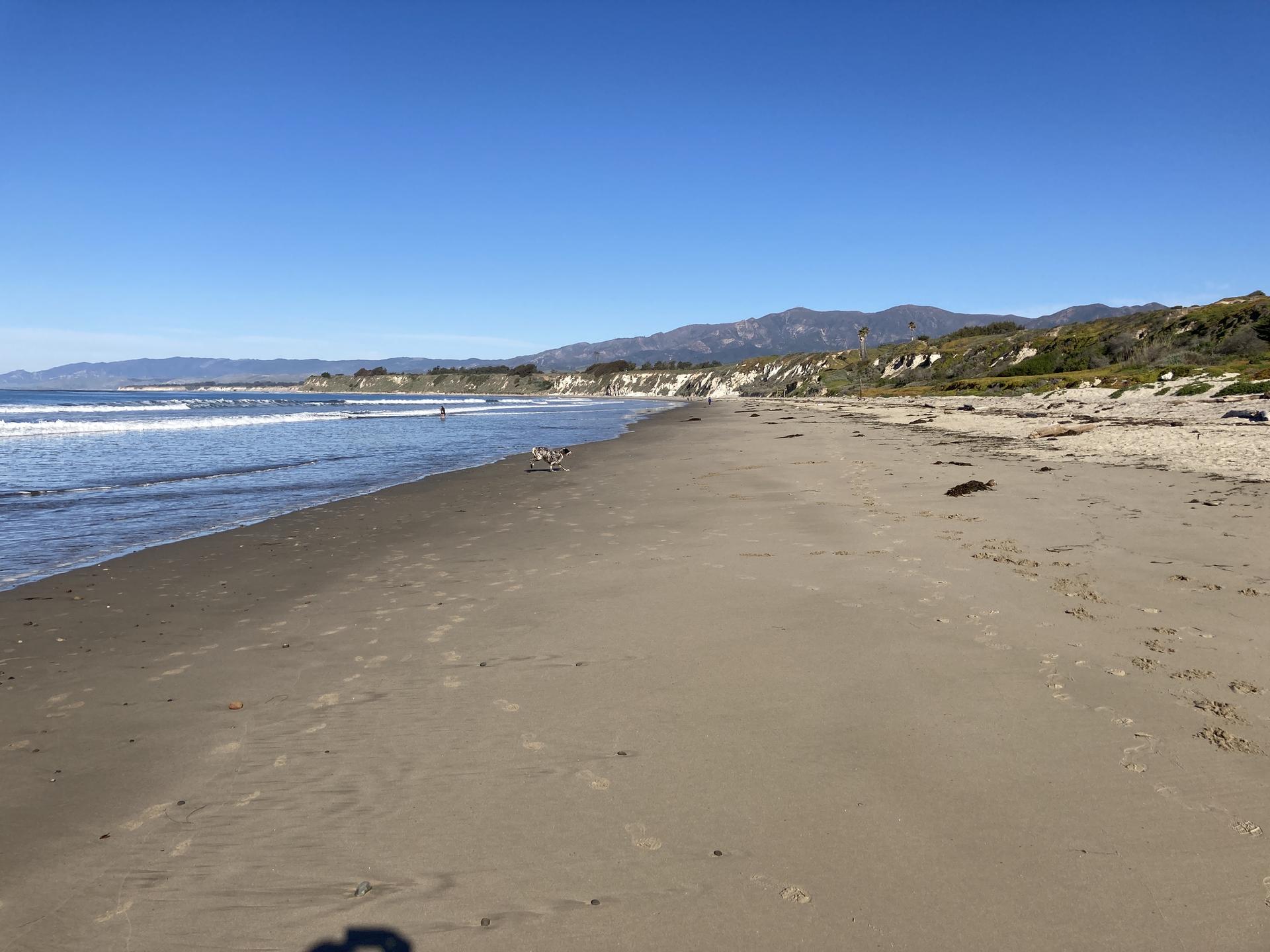 Expansive sandy beach with ocean waves and distant mountains under clear sky.