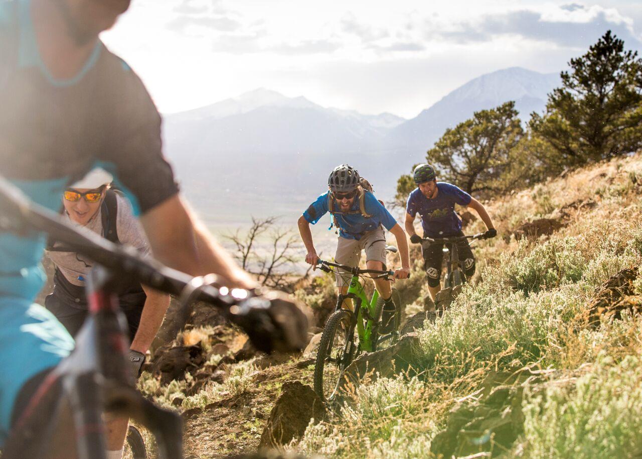 Three mountain bikers descend a sunny, dry, hilly trail with distant mountains.