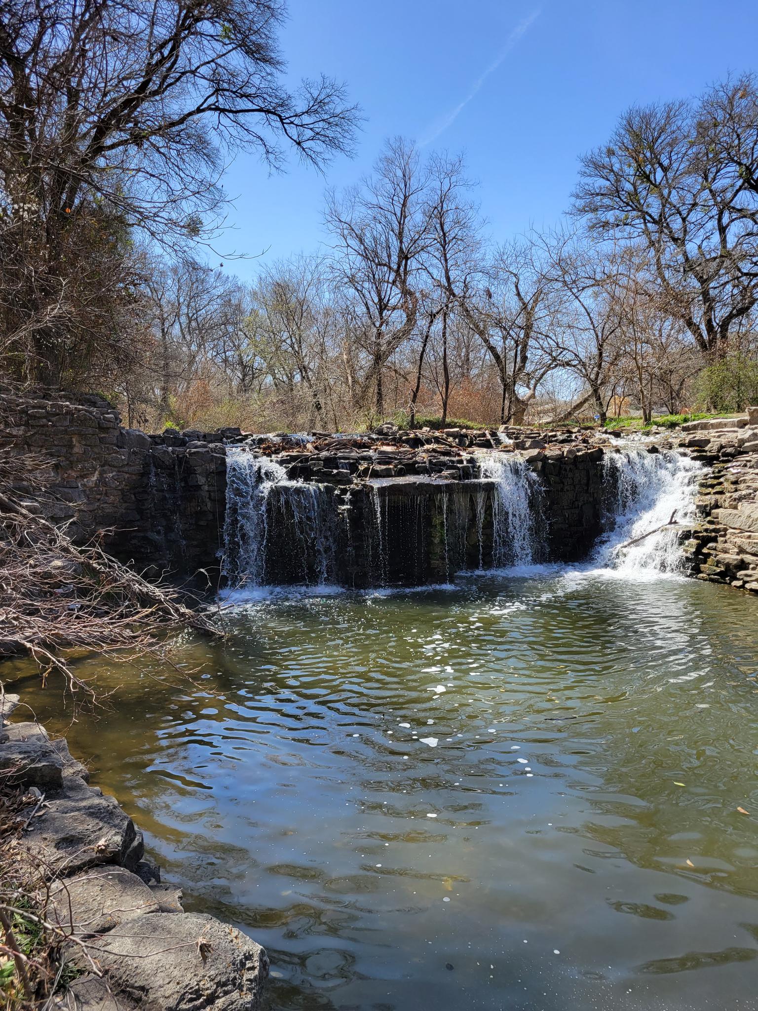 A multi-tiered waterfall cascades into a calm pool under a bright blue sky.