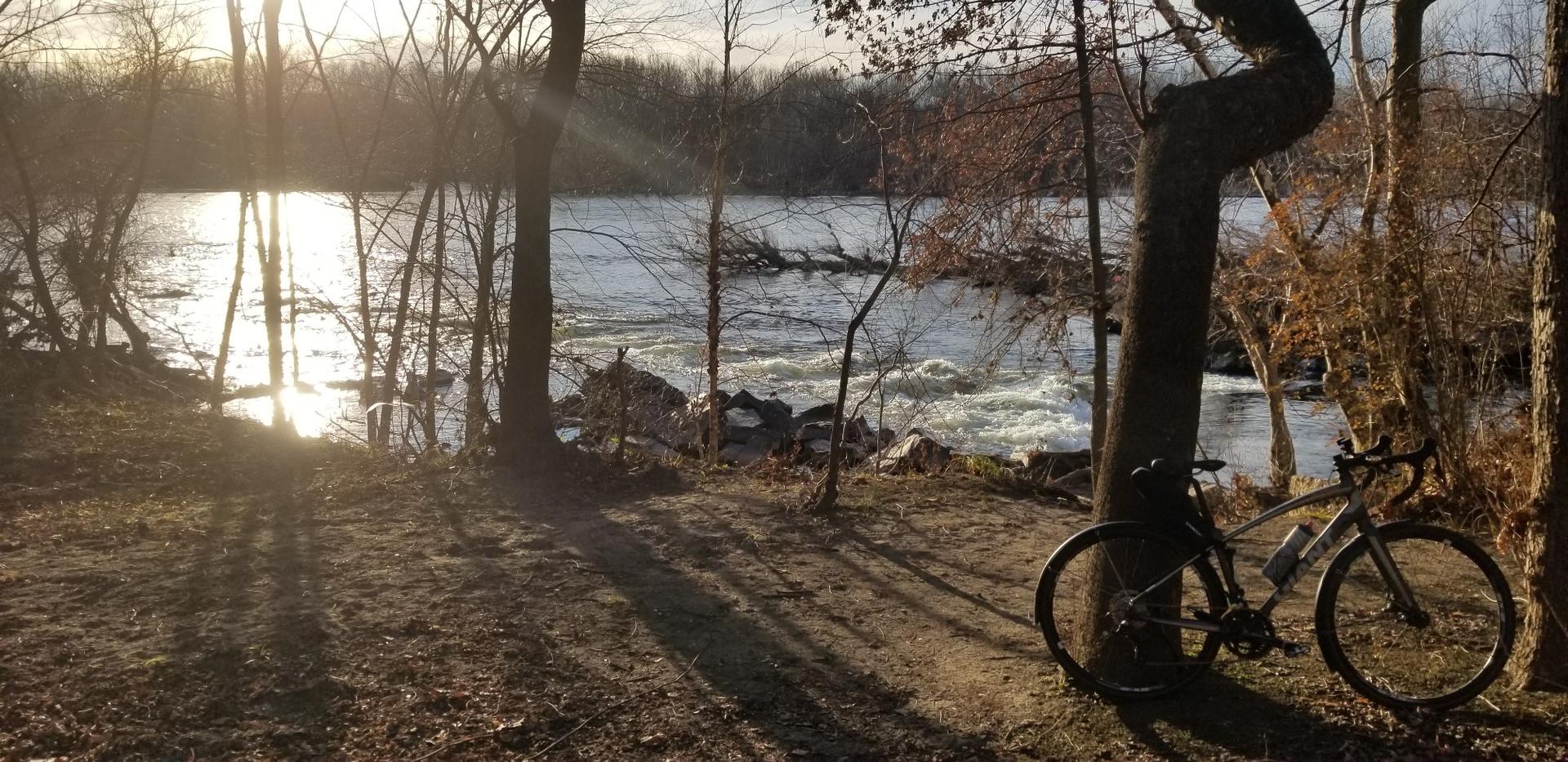 Bicycle by a river with rapids, sun setting through bare trees.
