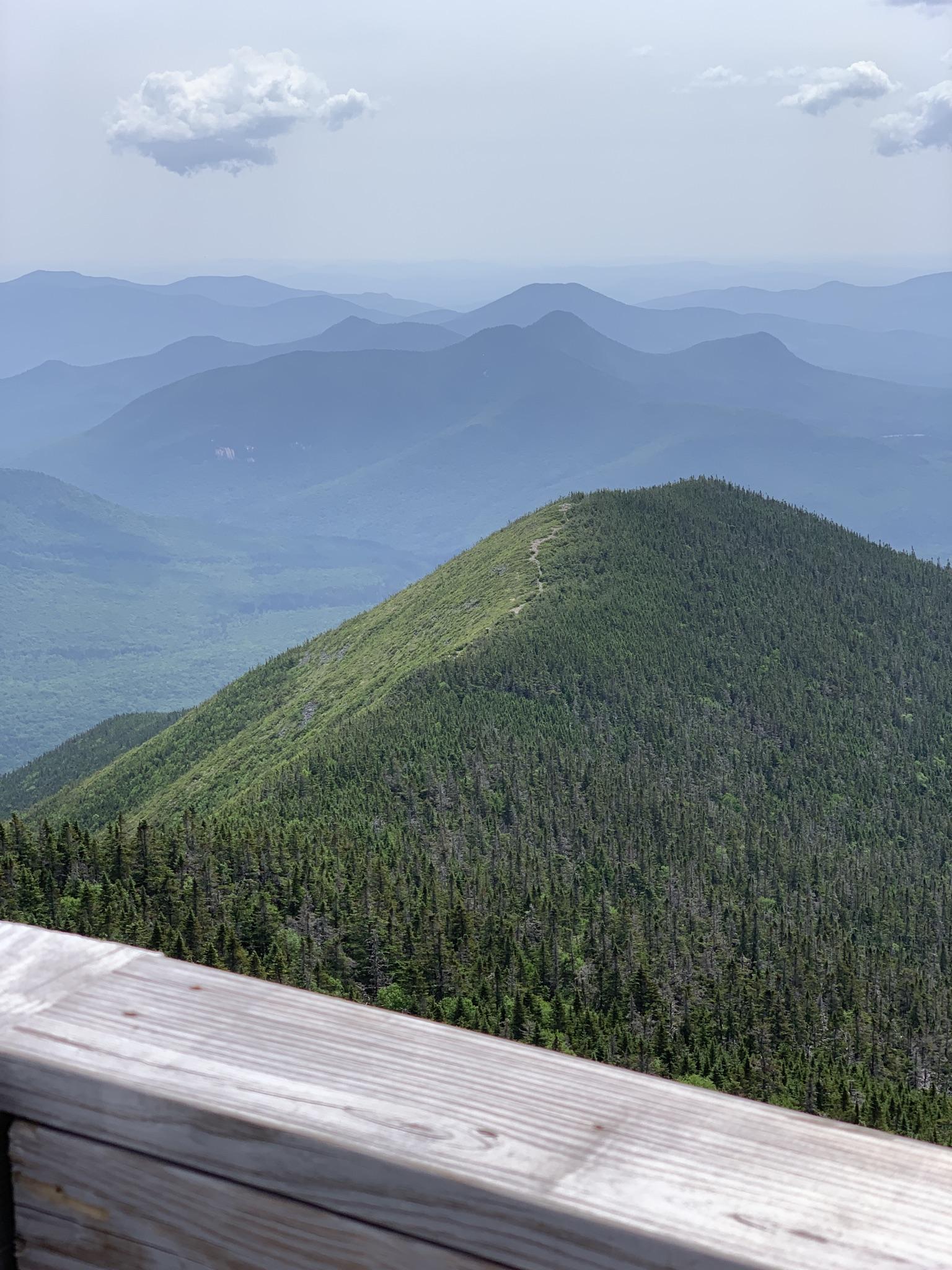 Forested mountain peak with distant hazy mountains under a bright, cloudy sky.