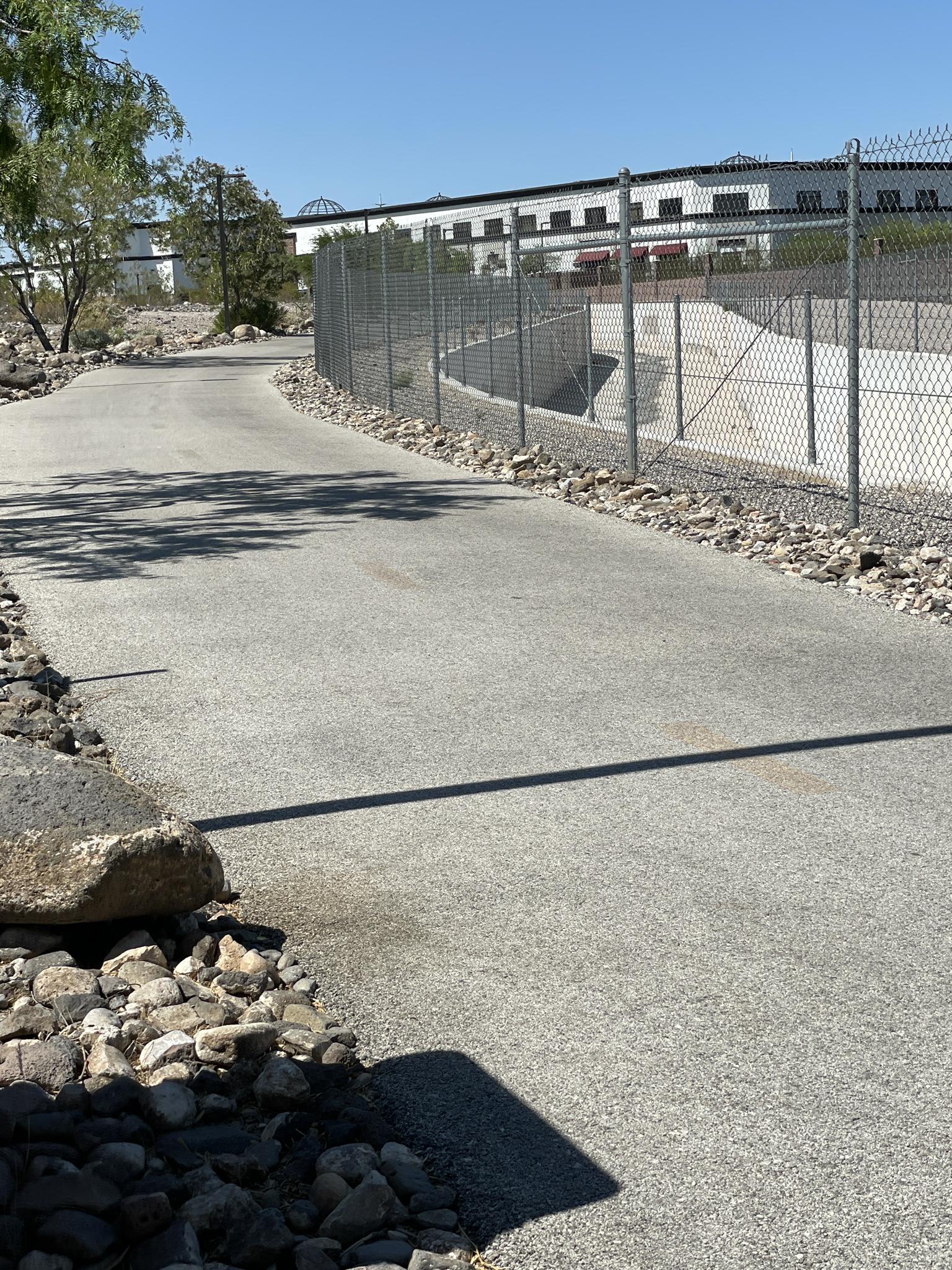 A paved path next to a chain-link fence and a large building.