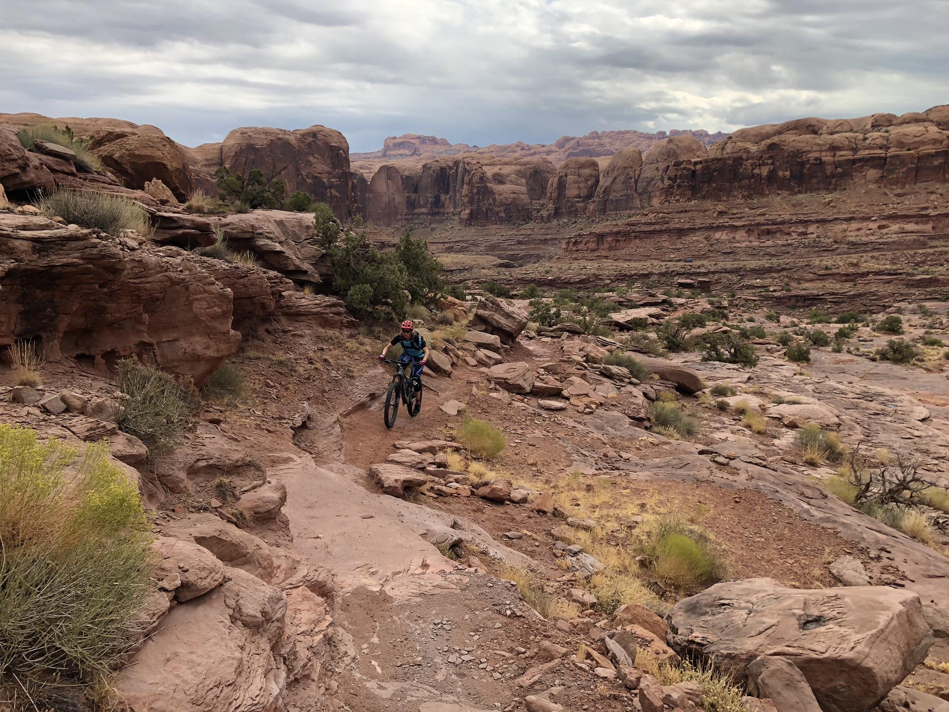 Mountain biker on a rocky trail in a wide canyon under a cloudy sky.