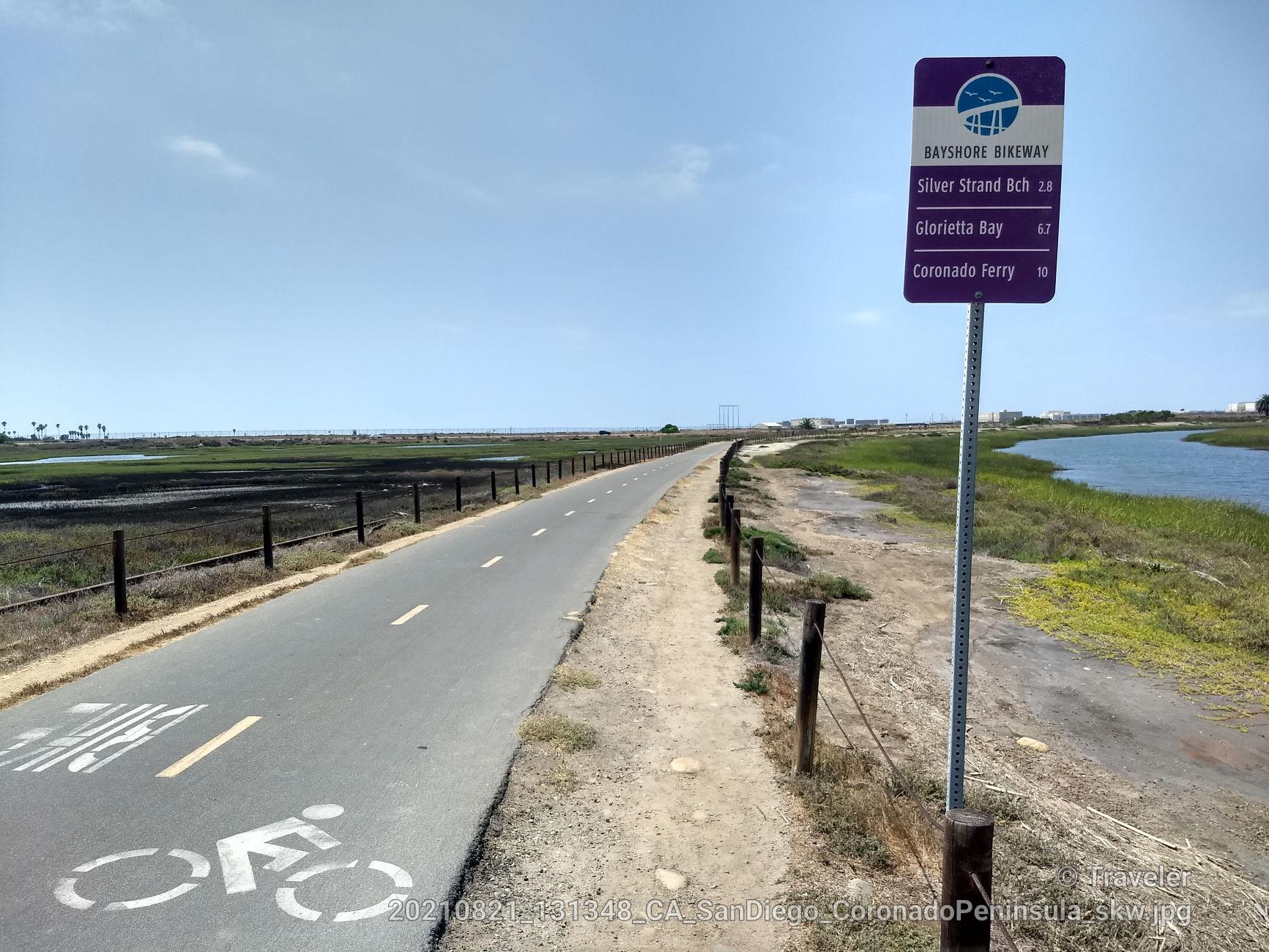 Paved bike path next to marshland and water under a clear sky.