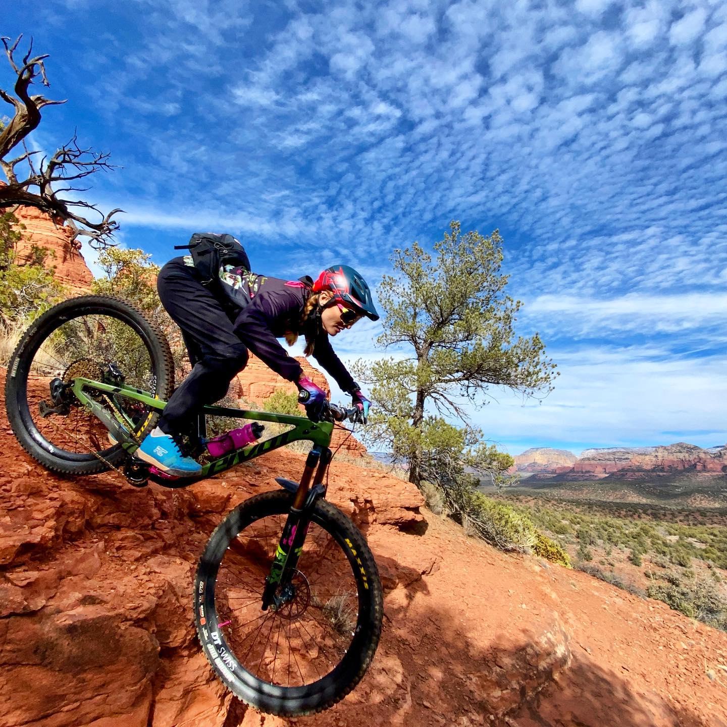 Mountain biker on a red rock ledge, bright blue sky with wispy clouds.
