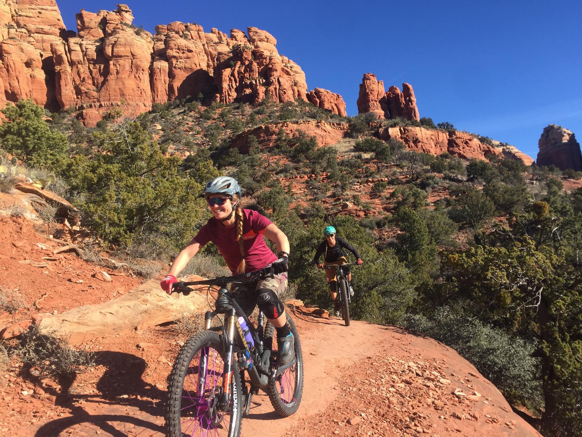 Two people mountain biking on a red dirt trail with large red rock formations.