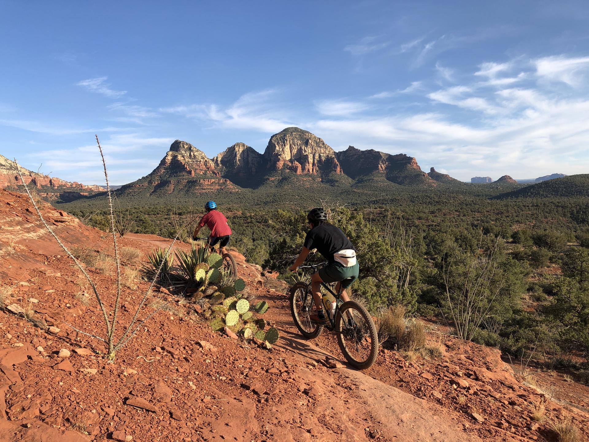 Two mountain bikers on a red dirt trail with distant red rock mountains.