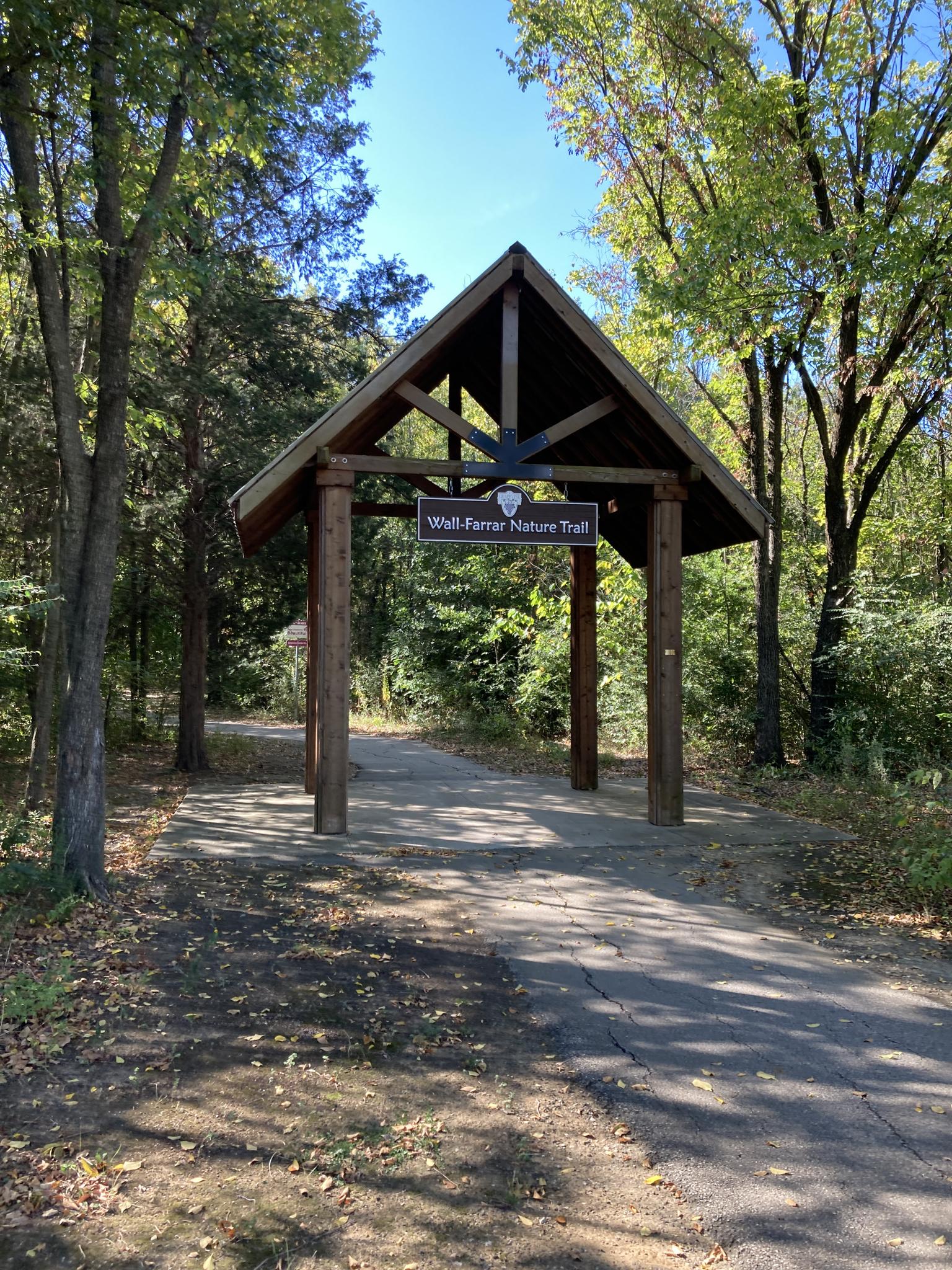 Wooden archway over a paved trail entrance, surrounded by green trees on a sunny day.