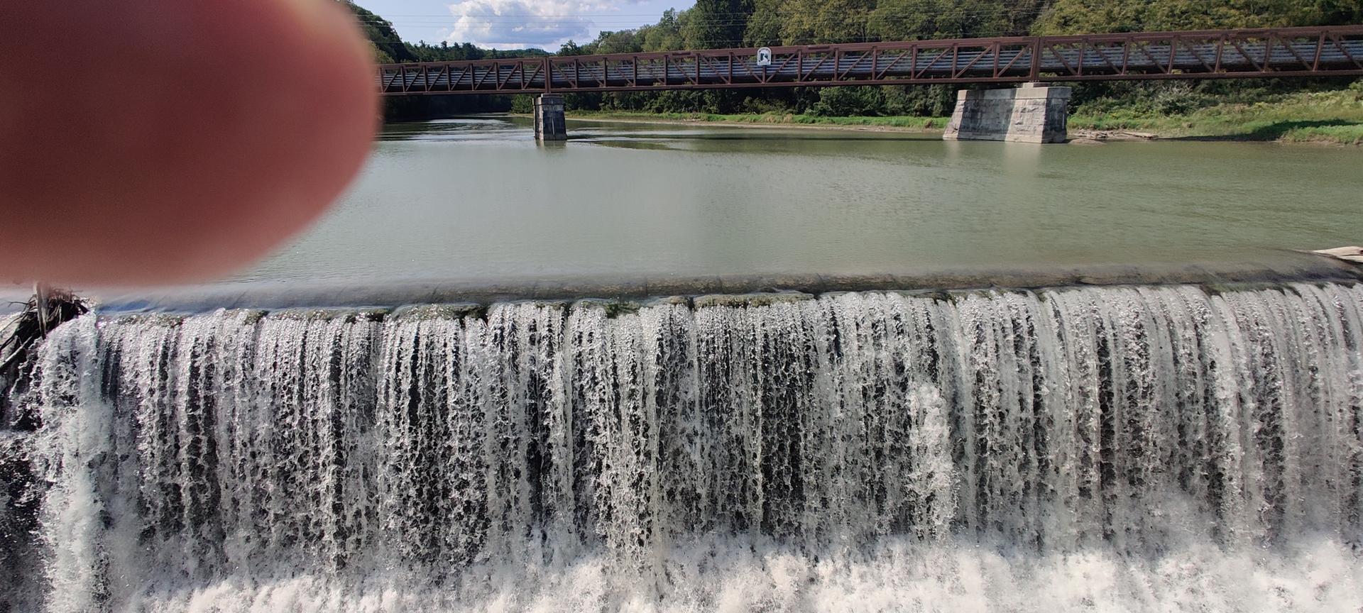 A wide waterfall cascades over a river with a bridge under bright daylight.