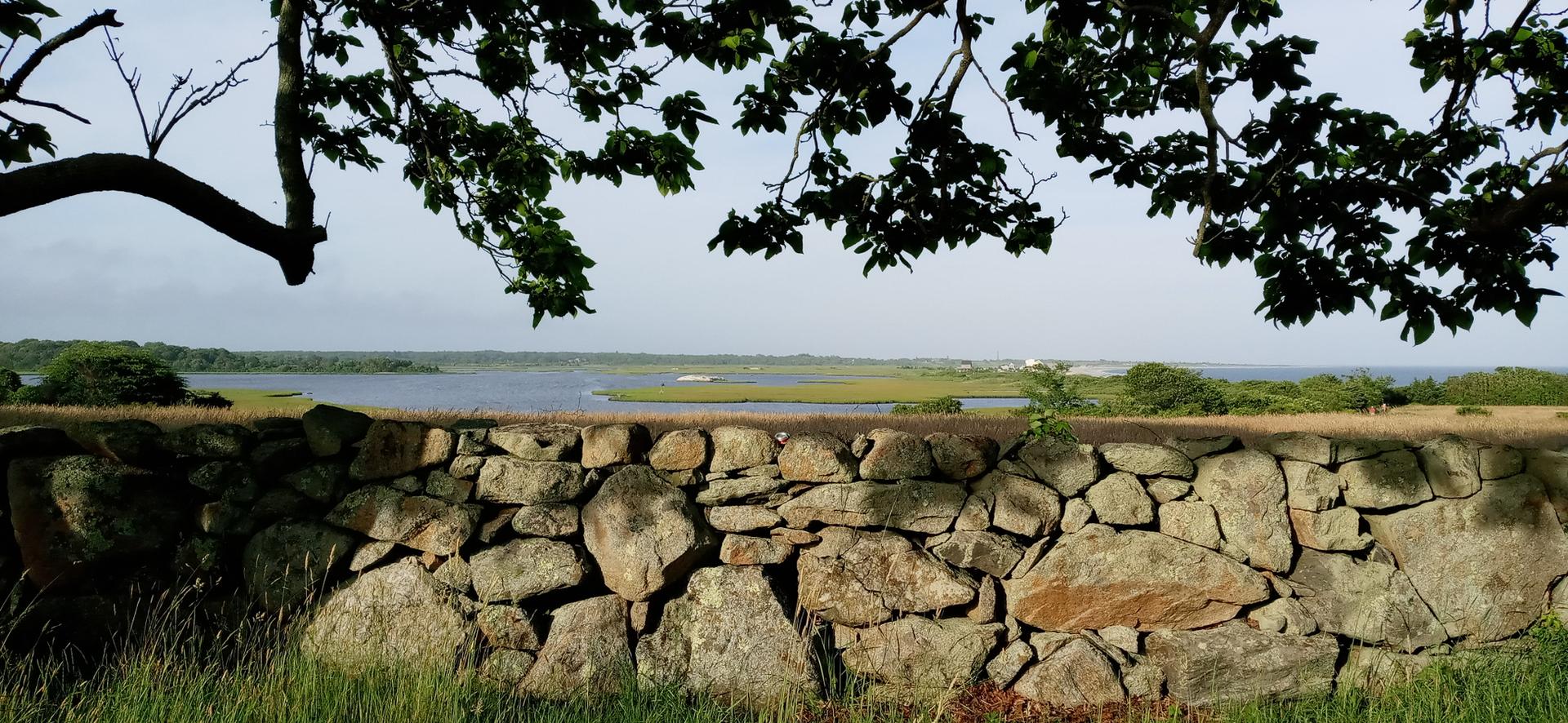 Stone wall in foreground, overlooking a wide river and distant marshland under a hazy sky.