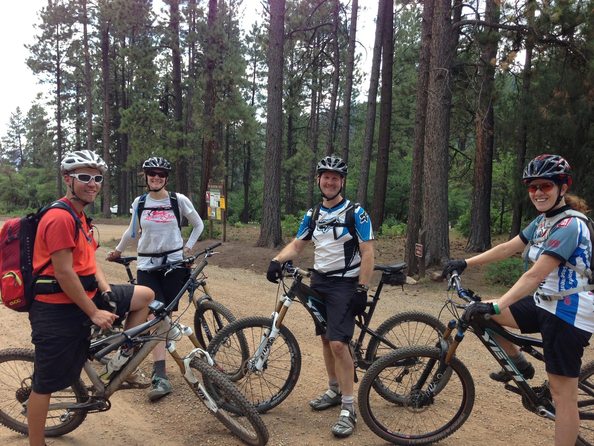 Four mountain bikers with helmets and bikes on a dirt path in a forest.