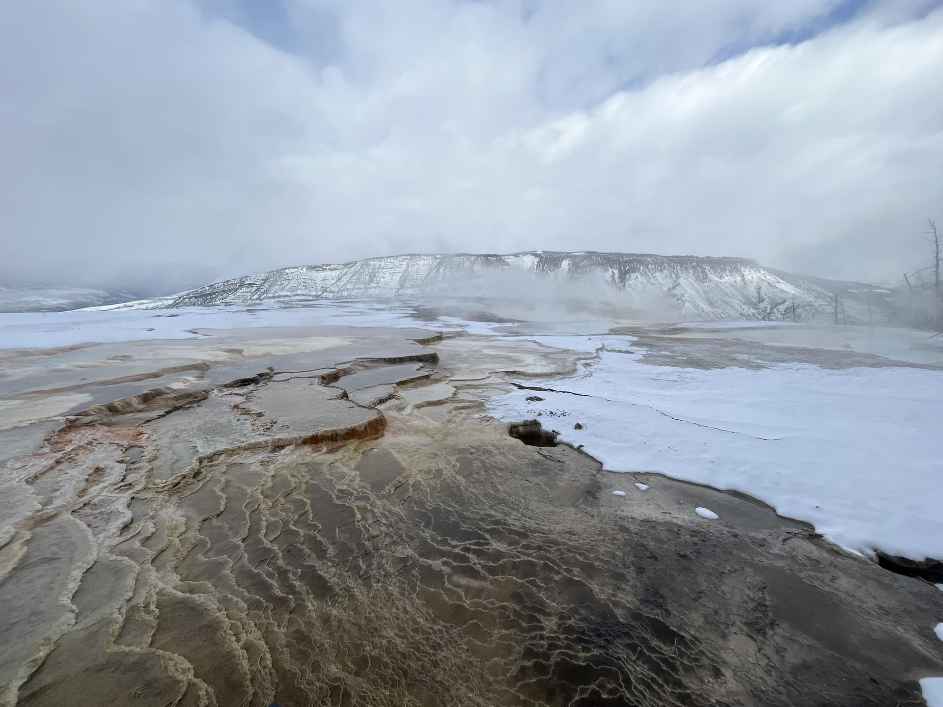 Steaming mineral formations and snow-covered ground with distant mountains under a cloudy sky.